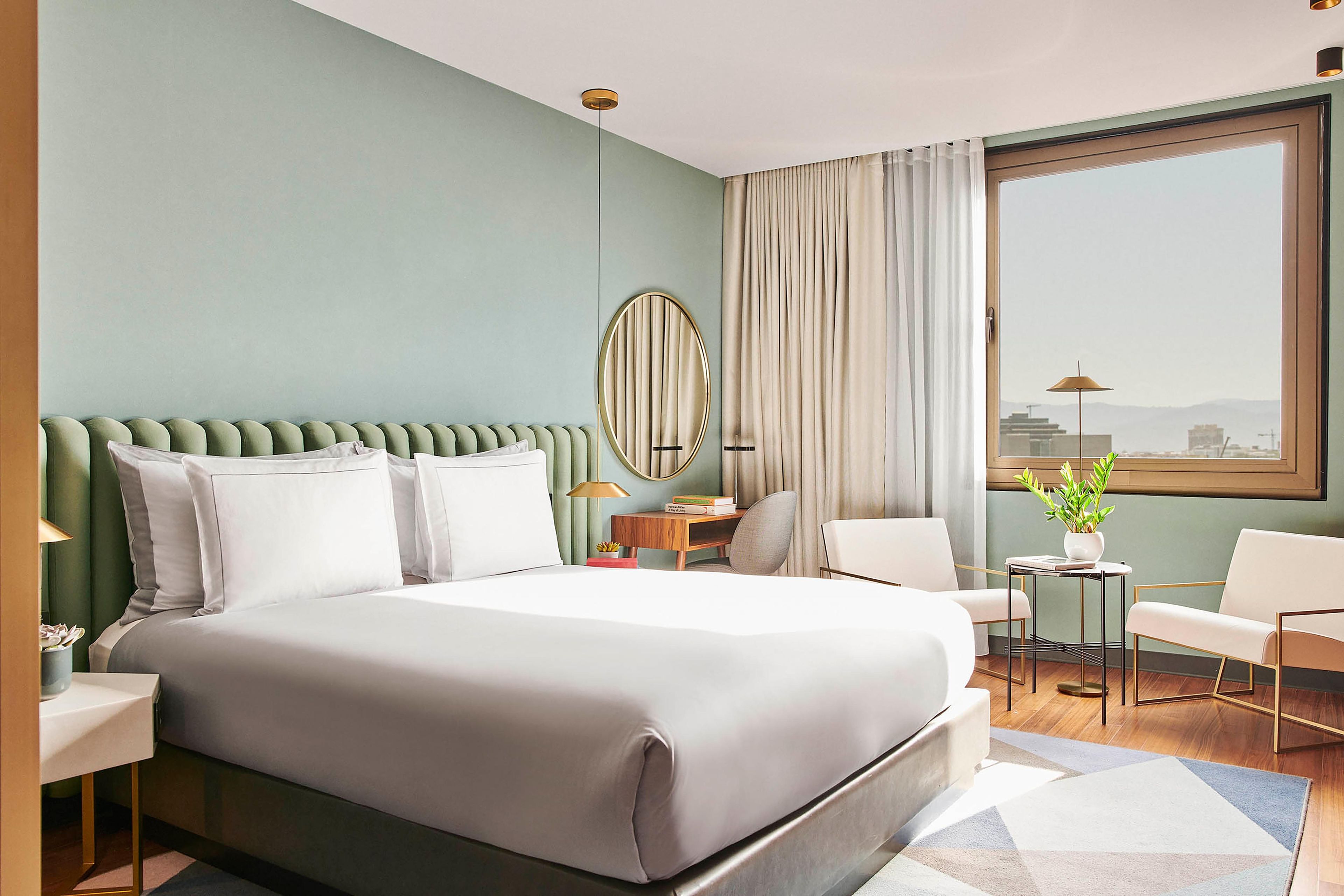 A hotel room with a large bed, a desk, two chairs, a window with a view, and a potted plant.