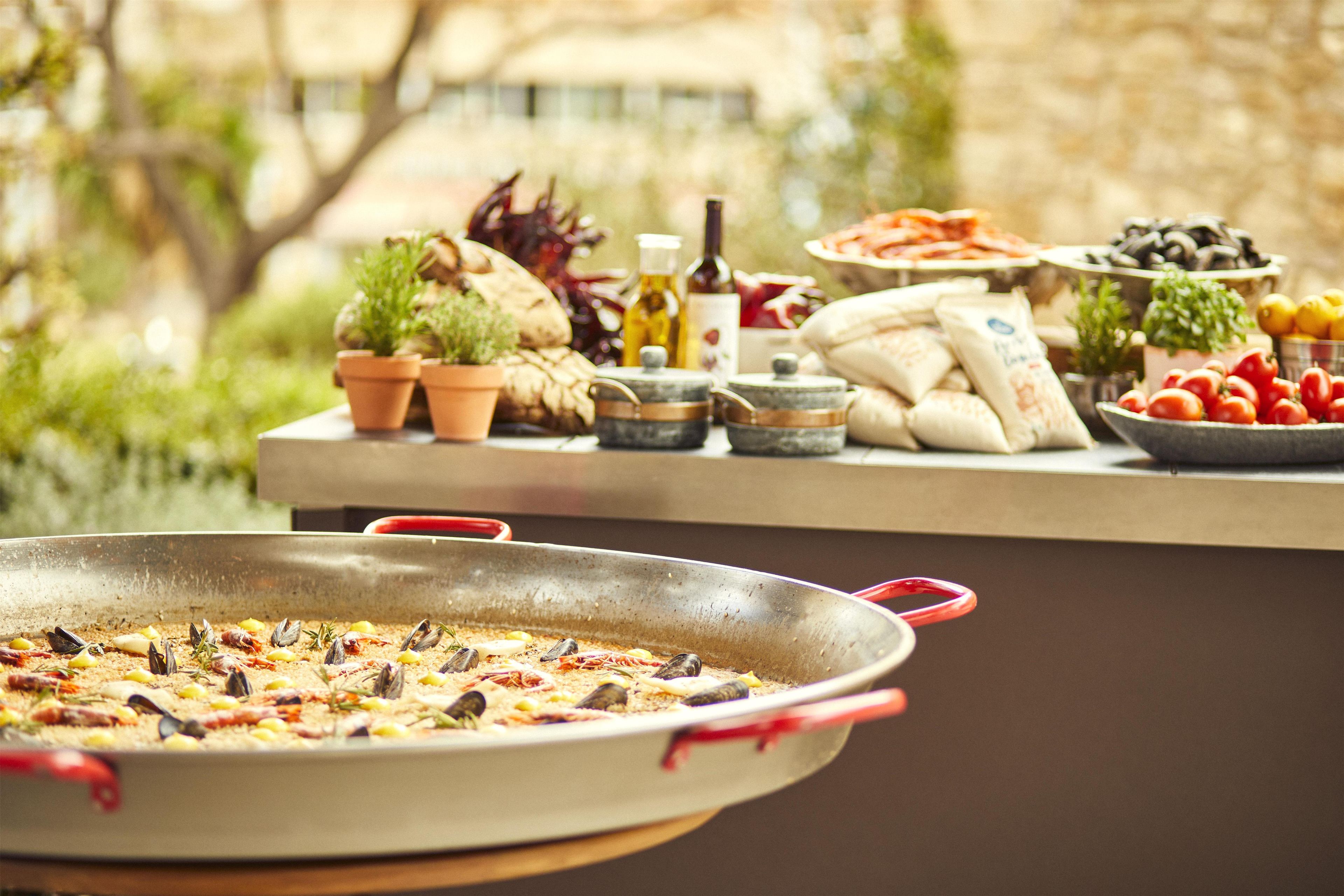 Una paella de mariscos en una sartén con mango rojo, varios platos que incluyen aceitunas y tomates, y una botella de aceite de oliva sobre una mesa.