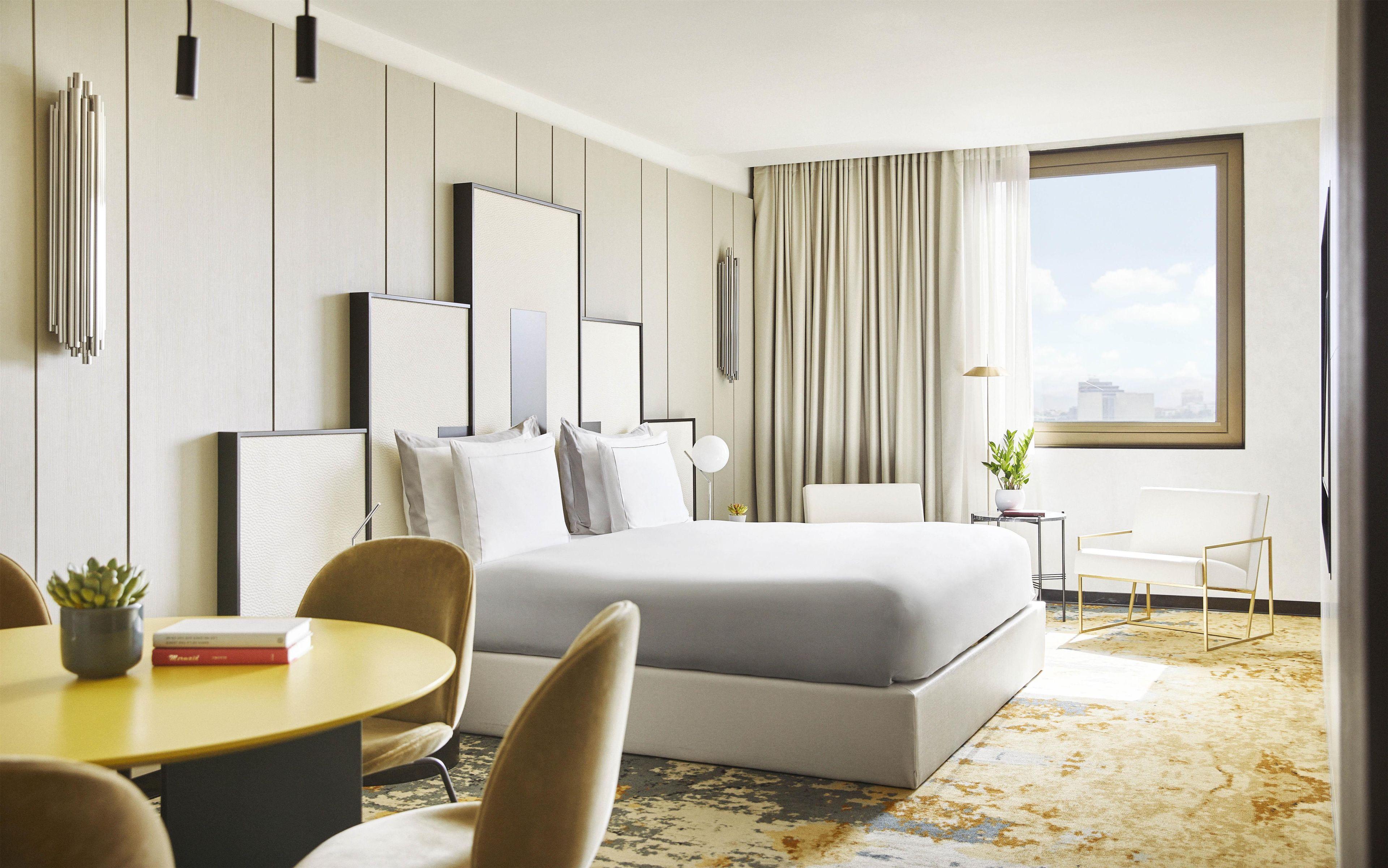 A modern hotel room with a large bed, a dining area with a table and chairs, a small side table with a plant, and a large window with curtains.