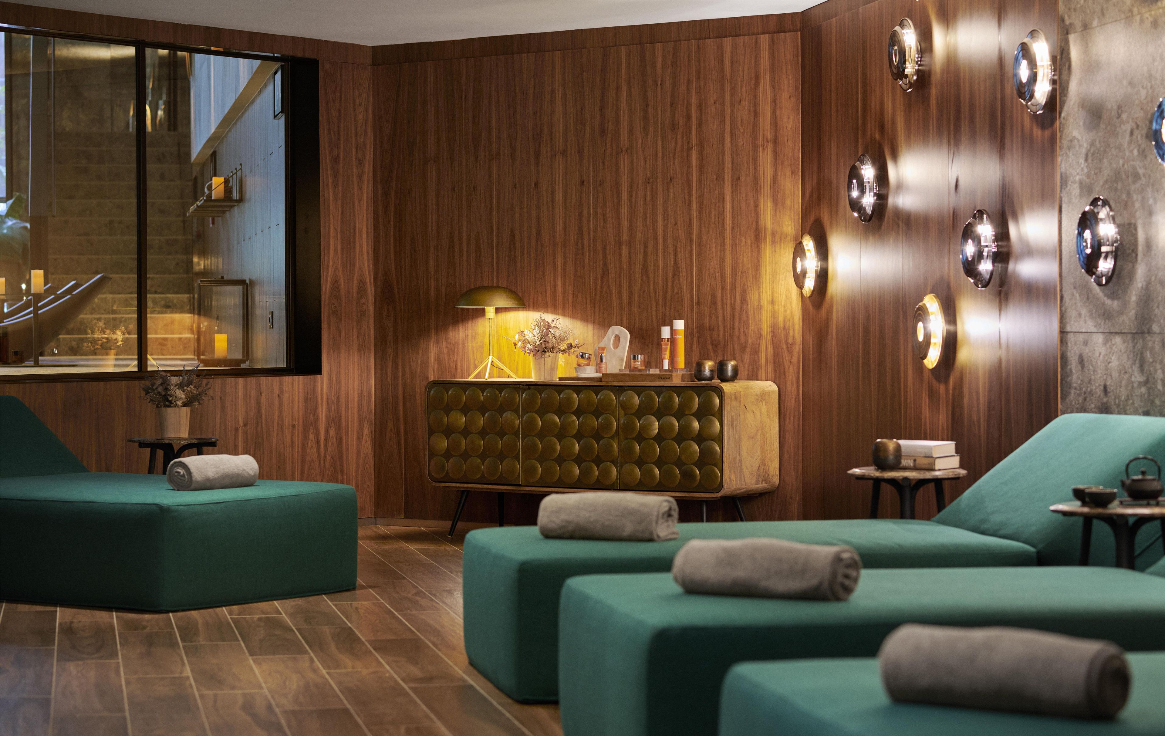 A modern interior with wooden panel walls, a green sofa set, a vintage-style console table, and a large window with a view of a staircase.