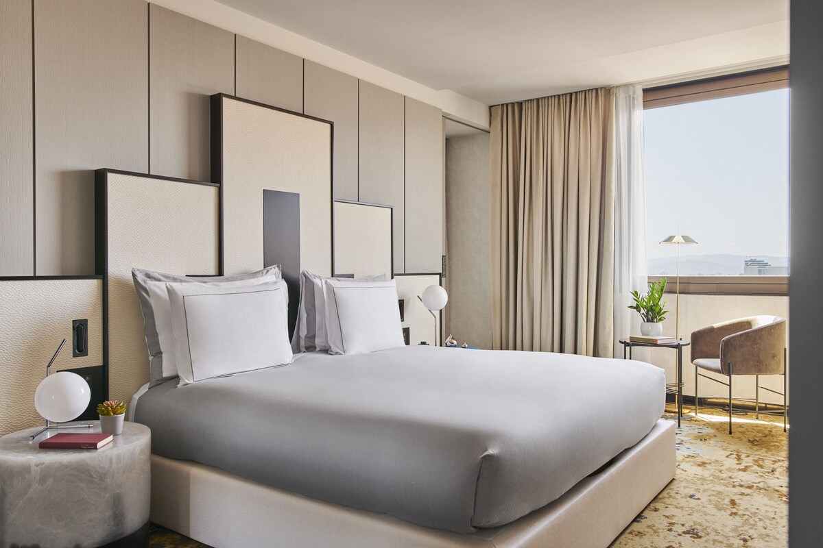 A modern hotel room with a large bed, a bedside table with a lamp, a chair, and a view of the city through the window.
