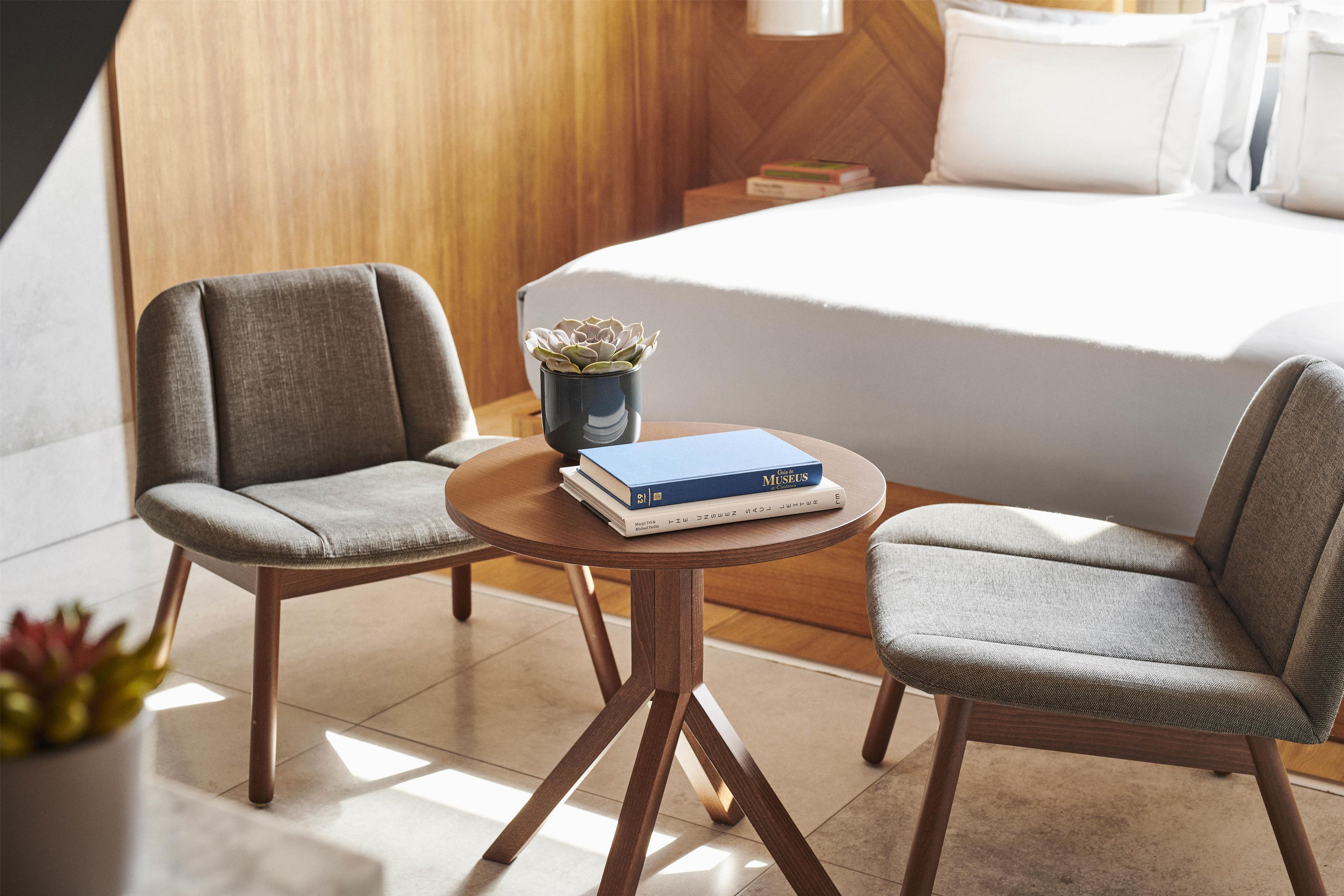 A hotel room with a bed, two armchairs, a small table with books, and a potted plant.