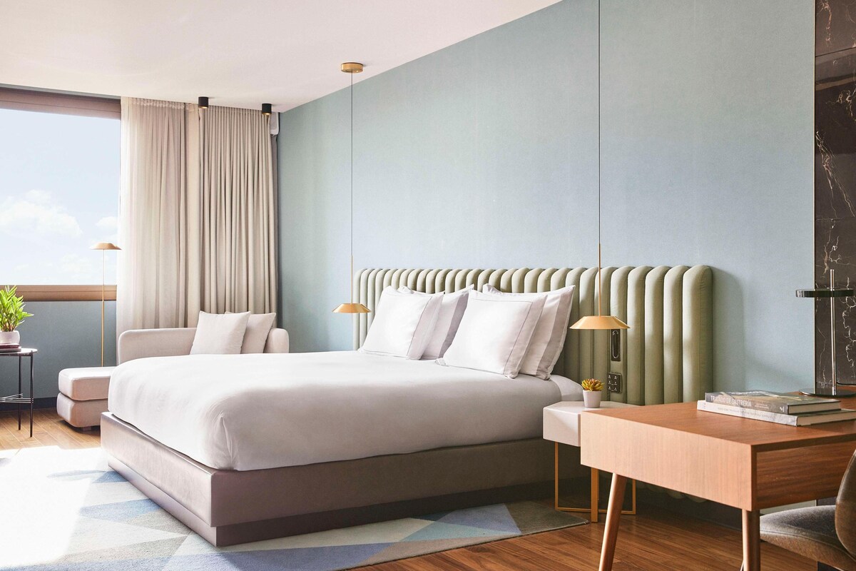 A modern hotel room with a large bed, a desk, and a view of the sky.