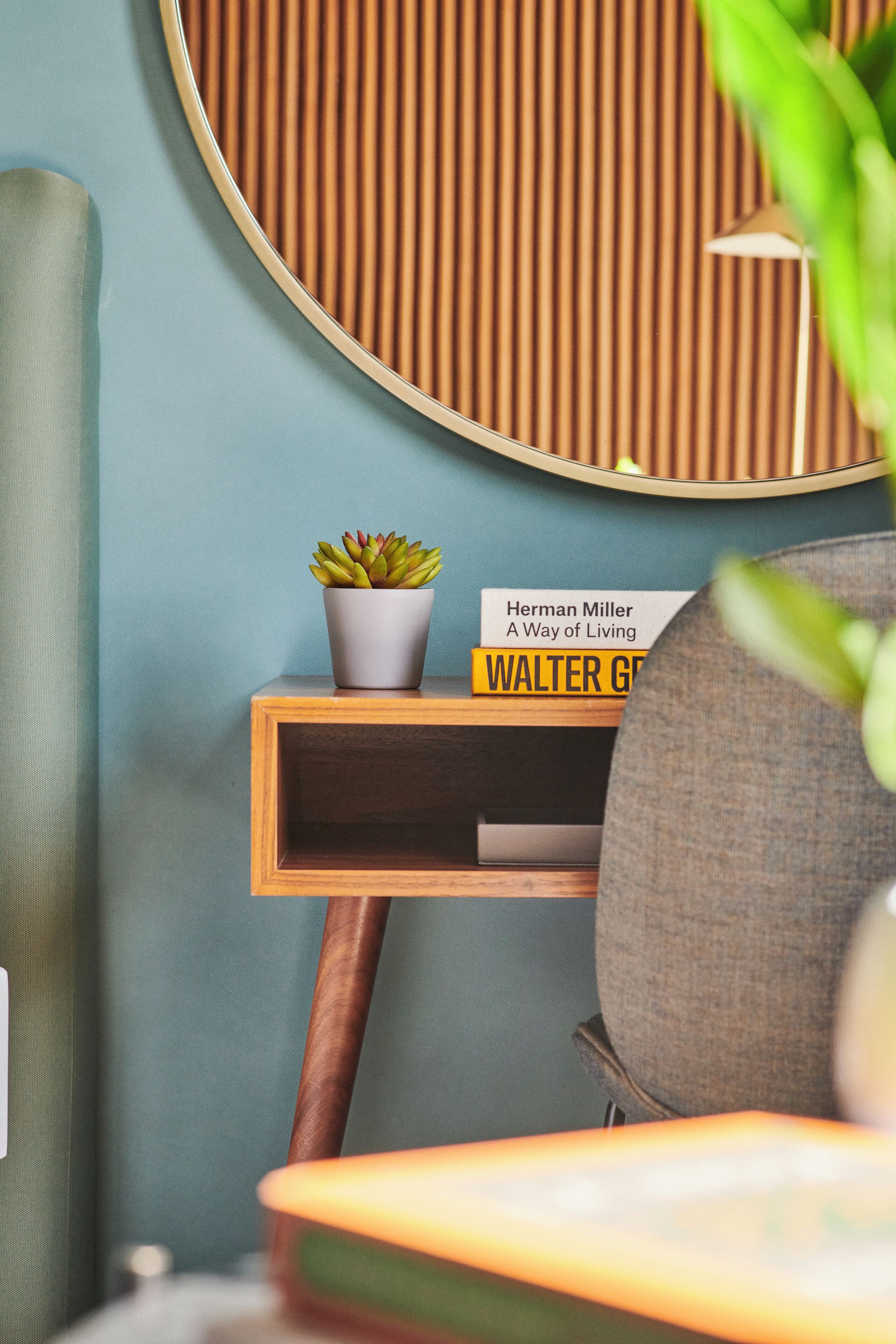 Une chambre moderne avec un grand miroir rond, une petite table d’appoint en bois sur laquelle est posée une plante en pot, un livre intitulé « Une façon de vivre » et un fauteuil gris.