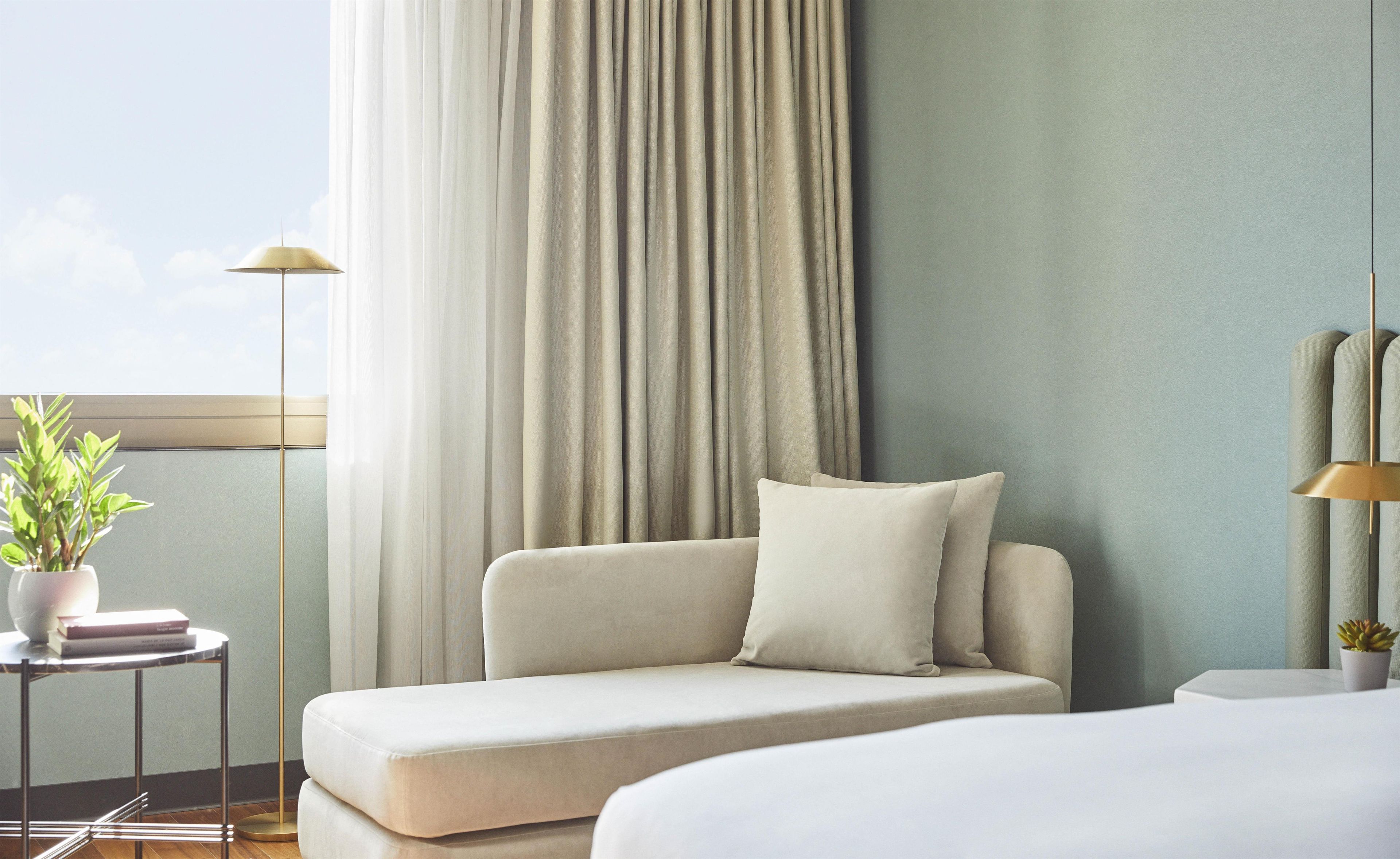 Quarto de hotel moderno com sofá, mesa de cabeceira iluminada por um abajur e janela com cortinas.