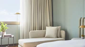 A modern hotel room with a sofa, a bedside table with a lamp, and a window with curtains.