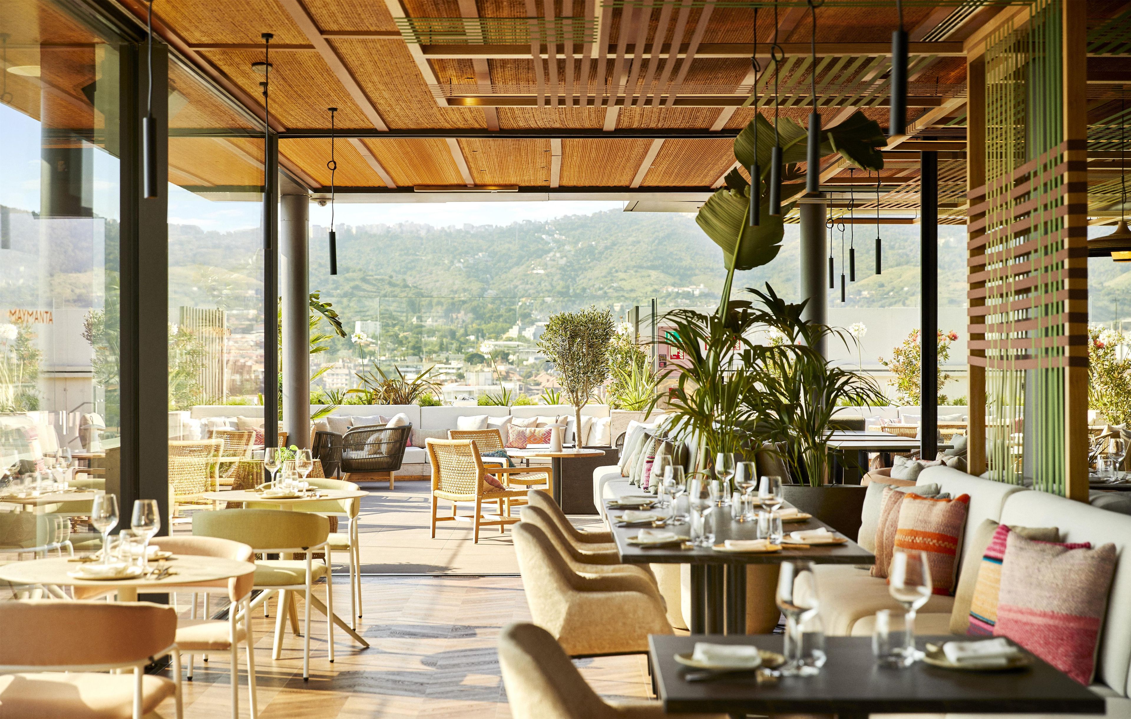 Un espacio exterior para comer con mesas y sillas, ventanas amplias y vistas a un paisaje montañoso.