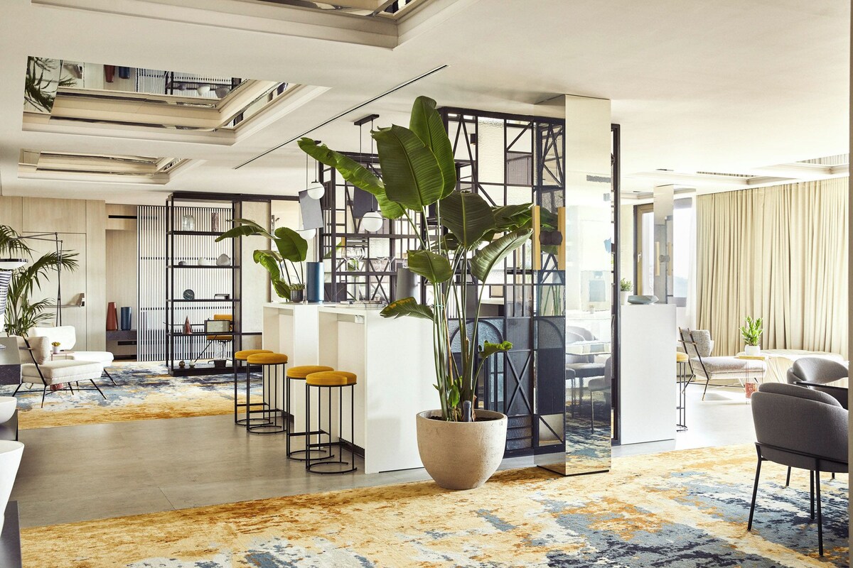 A modern office space with a large central plant, yellow bar stools, and a patterned carpet.
