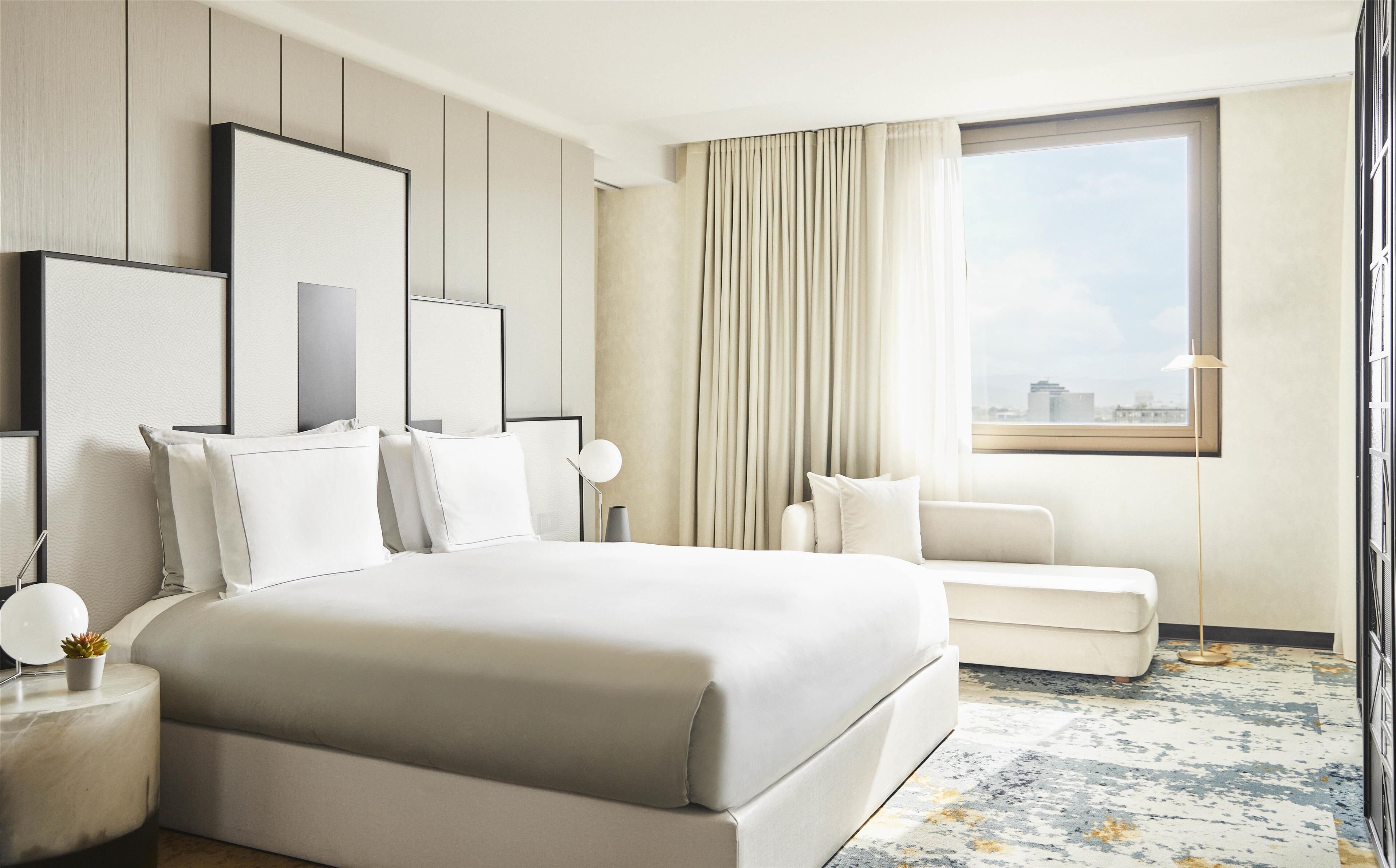 Una habitación de hotel moderna con una cama grande, zona de estar, vistas a la ciudad y un amplio ventanal con cortinas.