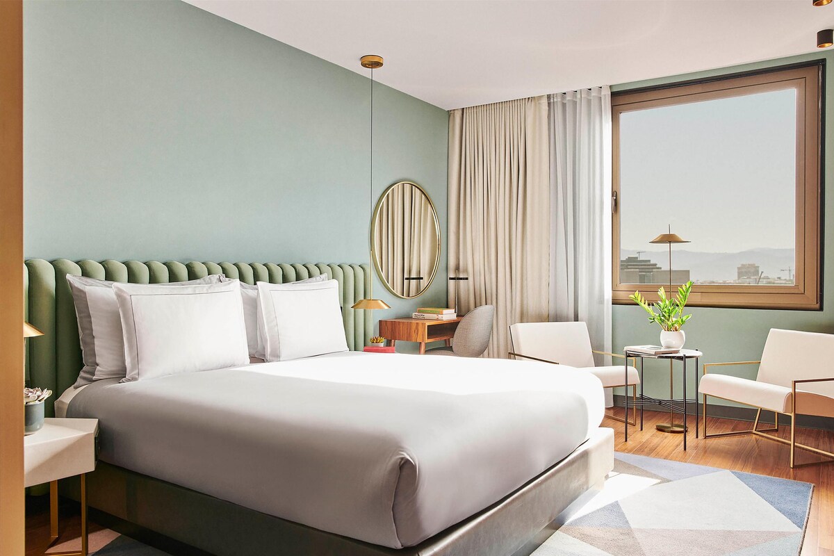 A modern hotel room with a large bed, a round mirror, a desk, two chairs, and a view of the city through a large window.