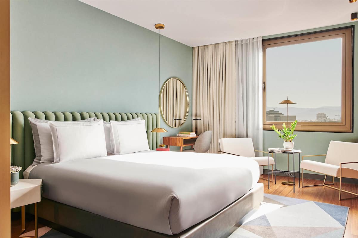 A modern hotel room with a large bed, a round mirror, a desk, two chairs, and a view of the city through a large window.