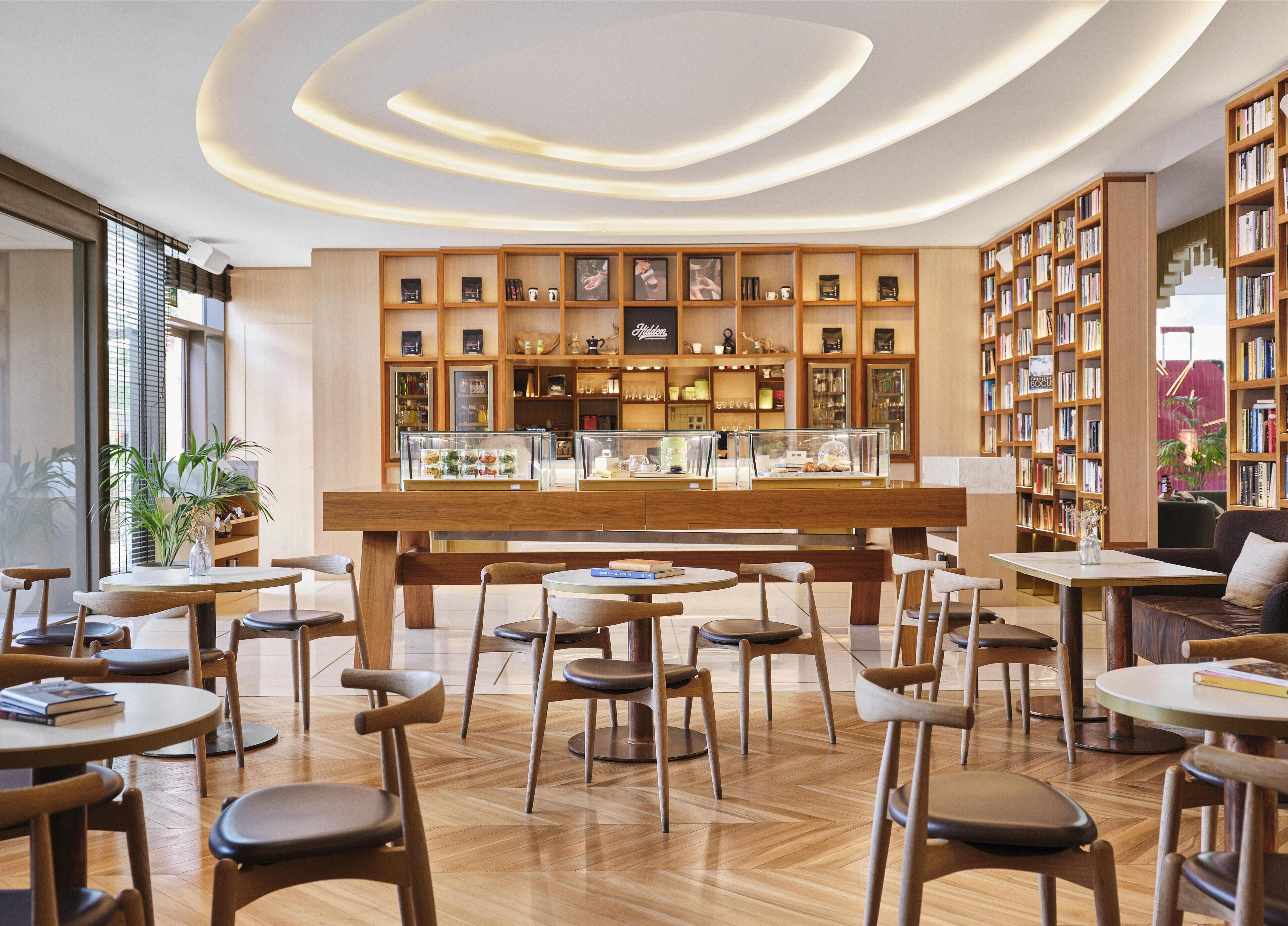 Un café moderno con mobiliario de madera, una barra central con vitrinas y estanterías llenas de libros.
