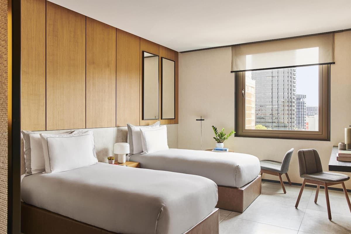 A hotel room with two beds, a desk, a chair, a potted plant, and a large window with a city view.