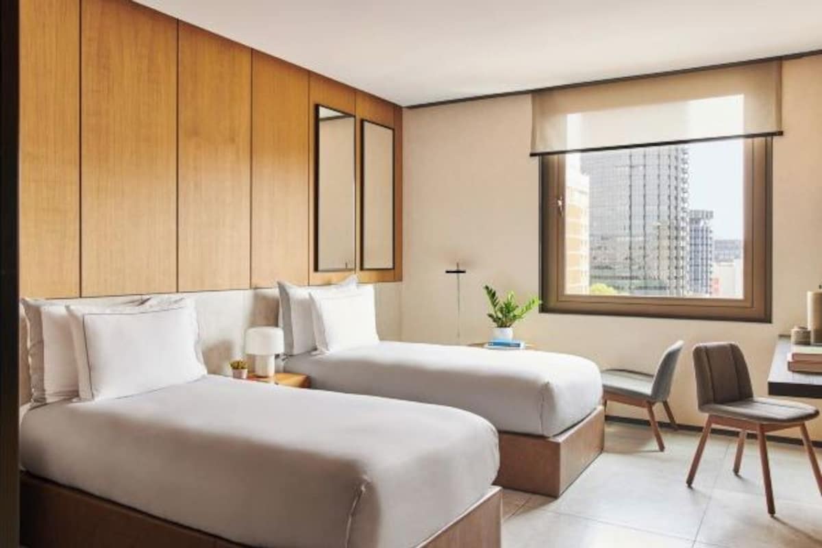 Une chambre d’hôtel avec deux lits, un bureau, une chaise, une grande fenêtre donnant sur la ville et un miroir au mur.