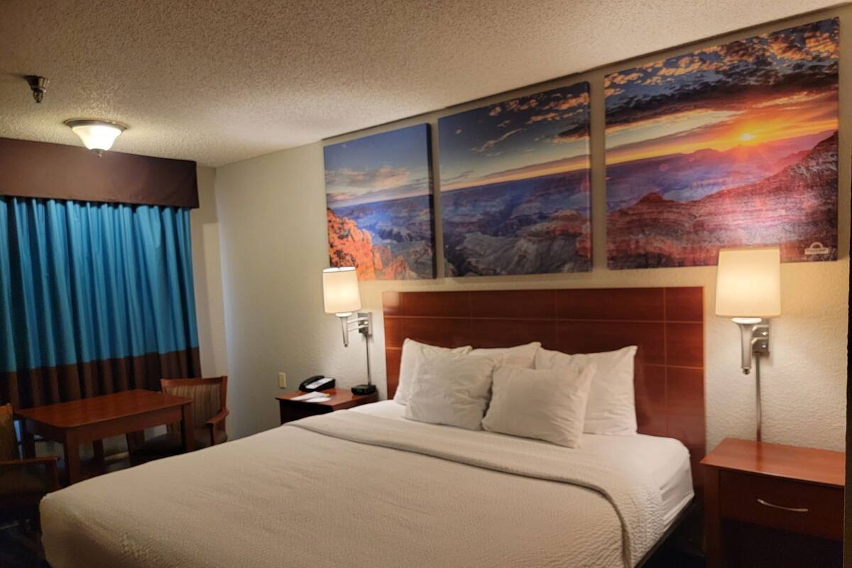 A hotel room with a large bed, a desk, two wall-mounted lamps, and a painting of a sunset over mountains.