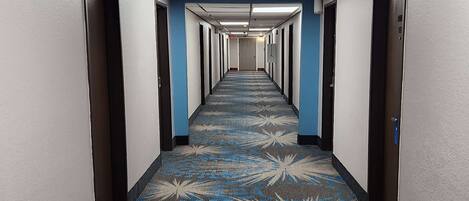 A hotel hallway with a patterned carpet, doors on both sides, and a ceiling with fluorescent lights.