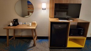 A hotel room with a wooden desk, a flat-screen TV, a mini-fridge, a microwave, a coffee maker, and a mirror.