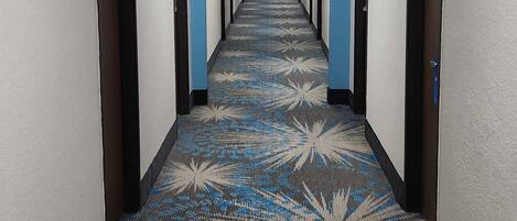 Un largo y estrecho pasillo de hotel con alfombra estampada, puertas a ambos lados y techo con iluminación fluorescente.