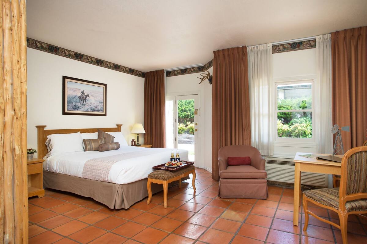 A hotel room with a bed, a desk, a chair, a window with curtains, a door, and a wall-mounted picture.