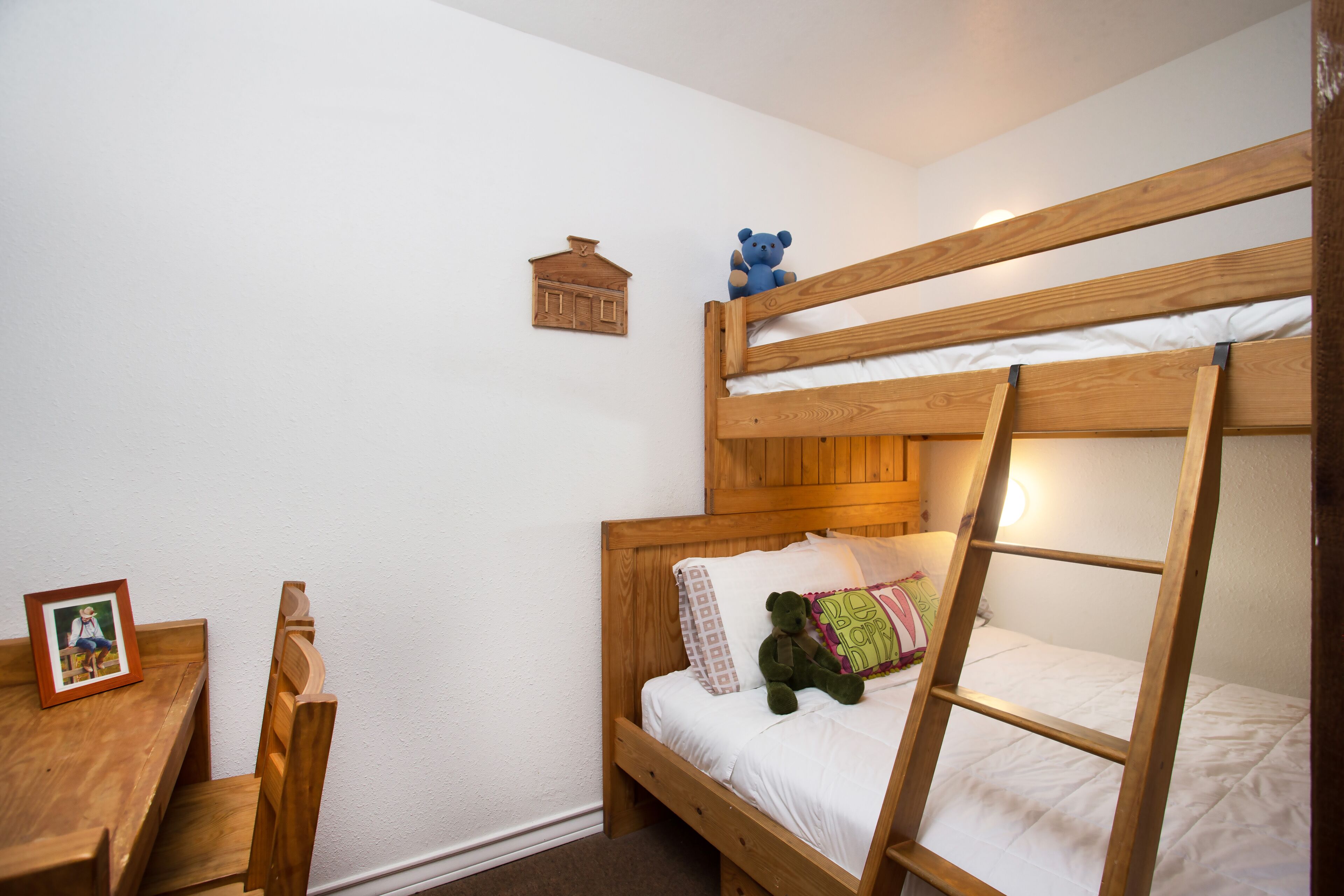 Una litera con escalera, un escritorio de madera, una silla, un oso de peluche y un adorno pequeño con forma de casa en la pared.
