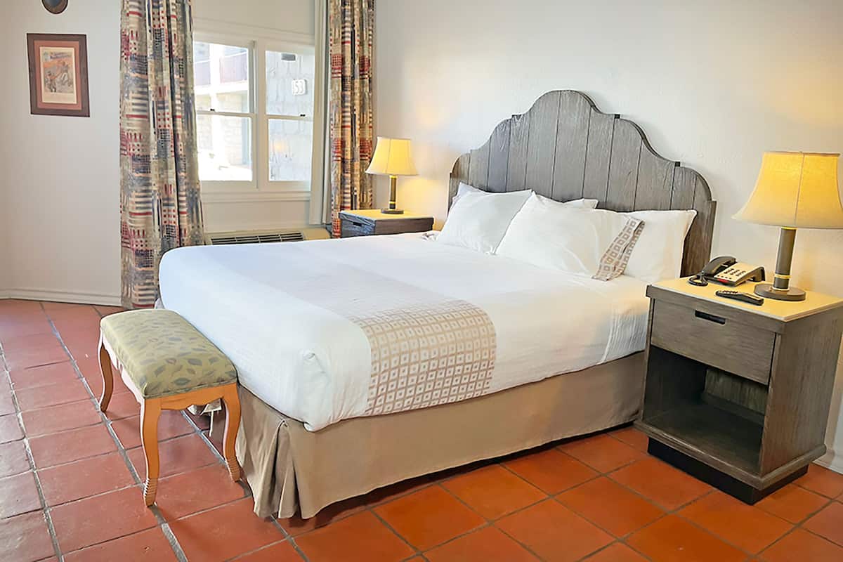 A hotel room with a large bed, two bedside tables with lamps, a bench, a window with curtains, and a wall-mounted deer head decoration.