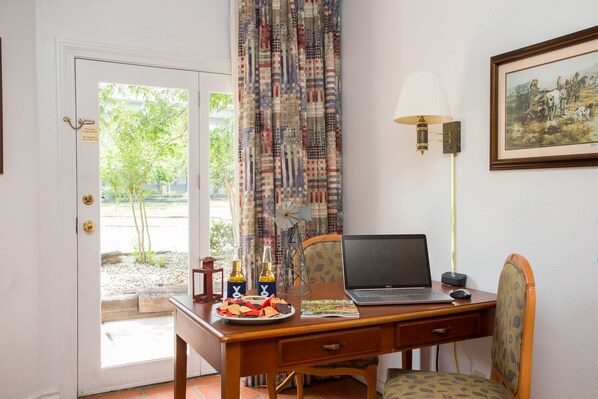 Una habitación con un escritorio de madera, una computadora portátil, una lámpara y una silla. Hay una puerta que da a un espacio al aire libre con árboles.