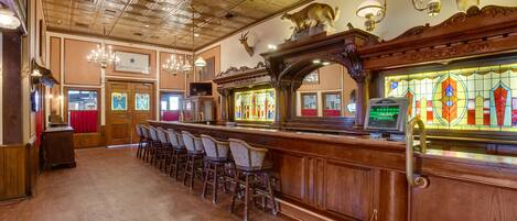 A bar area with wooden bar counters and chairs, a stained glass window, and a chandelier.