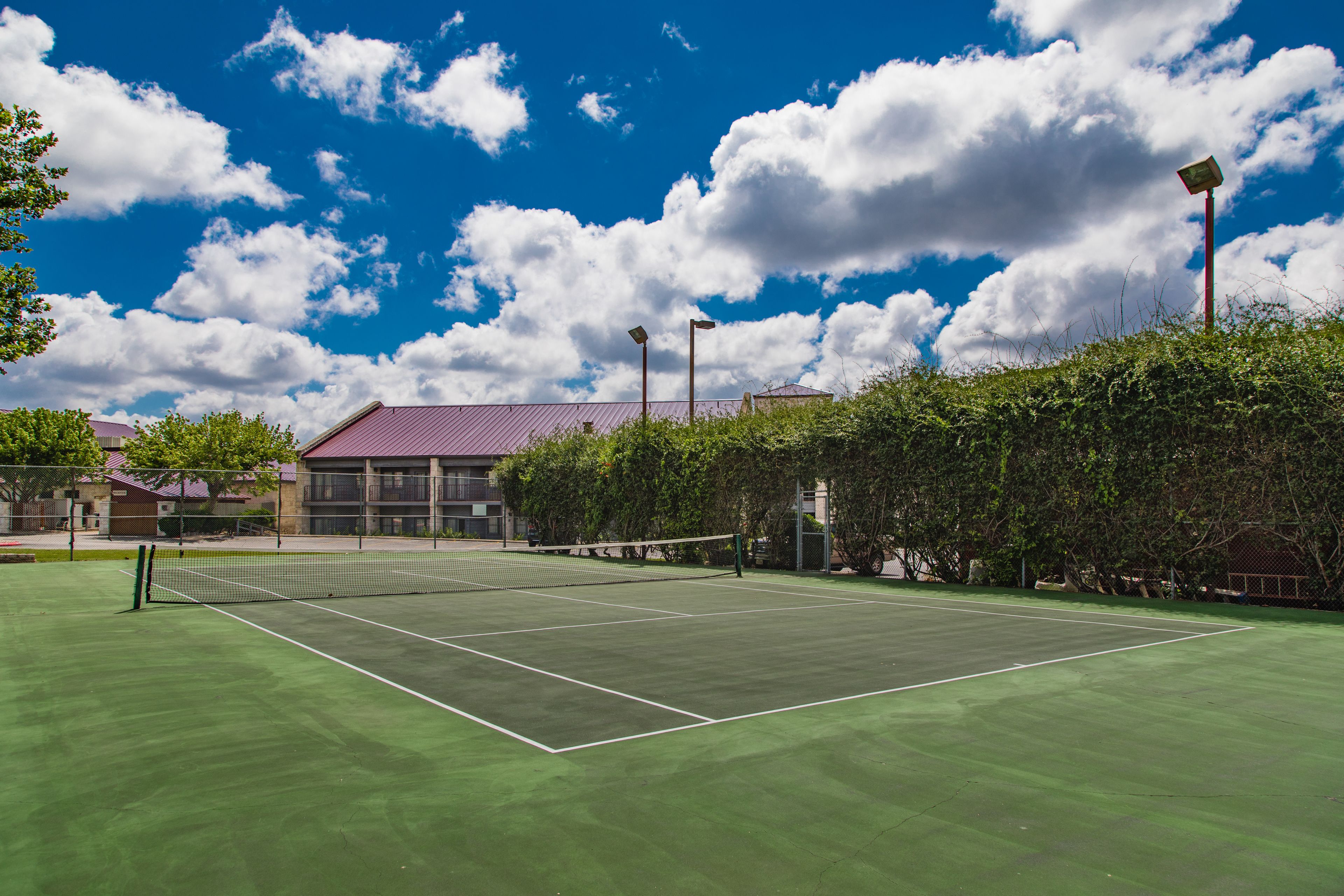 A tennis court with a net, surrounded by greenery and buildings in the background.