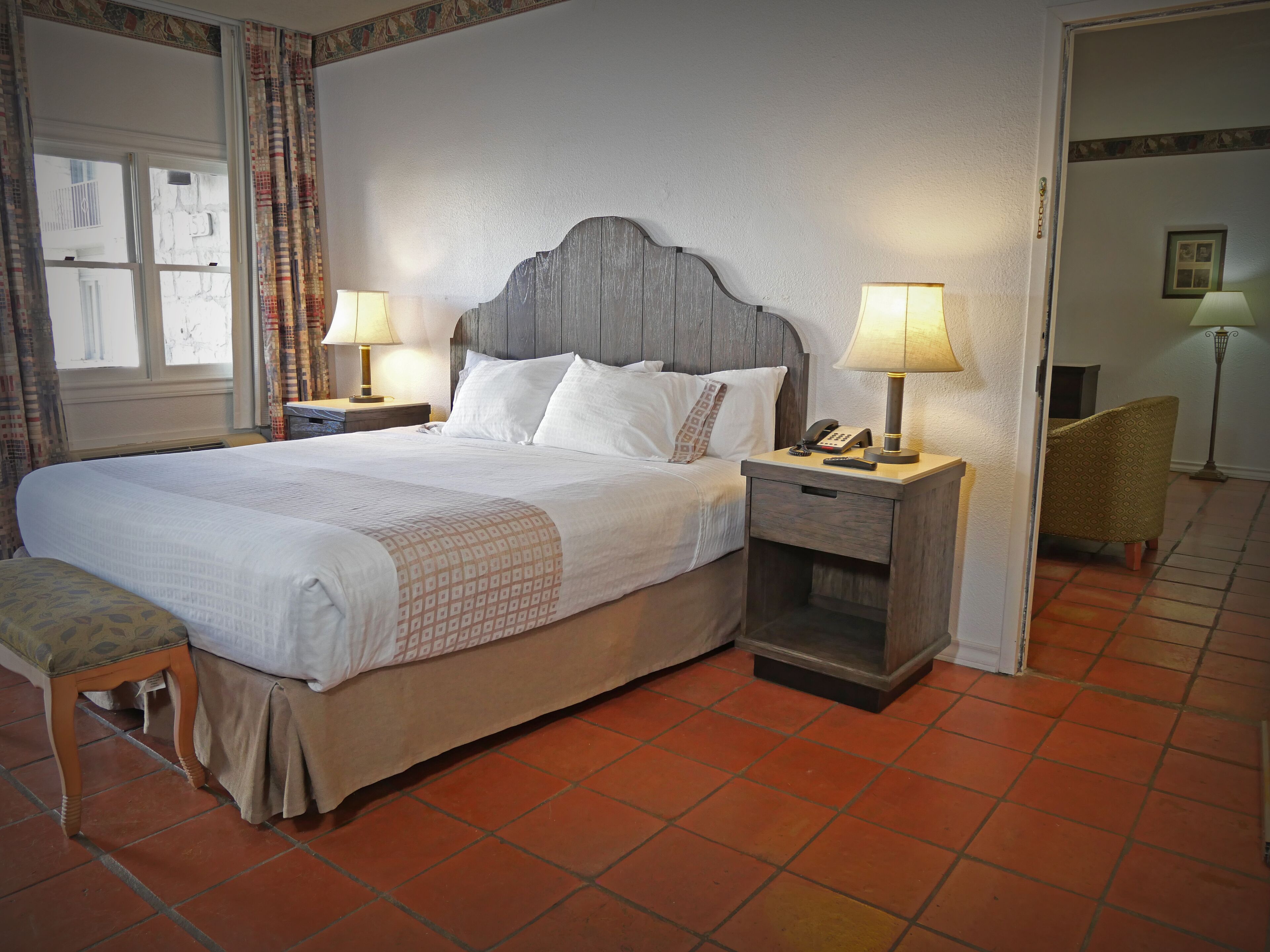 Un lit bien fait, avec du linge de lit blanc et un couvre-lit à motifs, une table de chevet avec une lampe, une chaise et une petite table sur laquelle se trouvent un téléphone et des télécommandes.