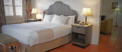 A neatly made bed with white linens and a patterned throw, a bedside table with a lamp, a chair, and a small table with a telephone and remote controls.