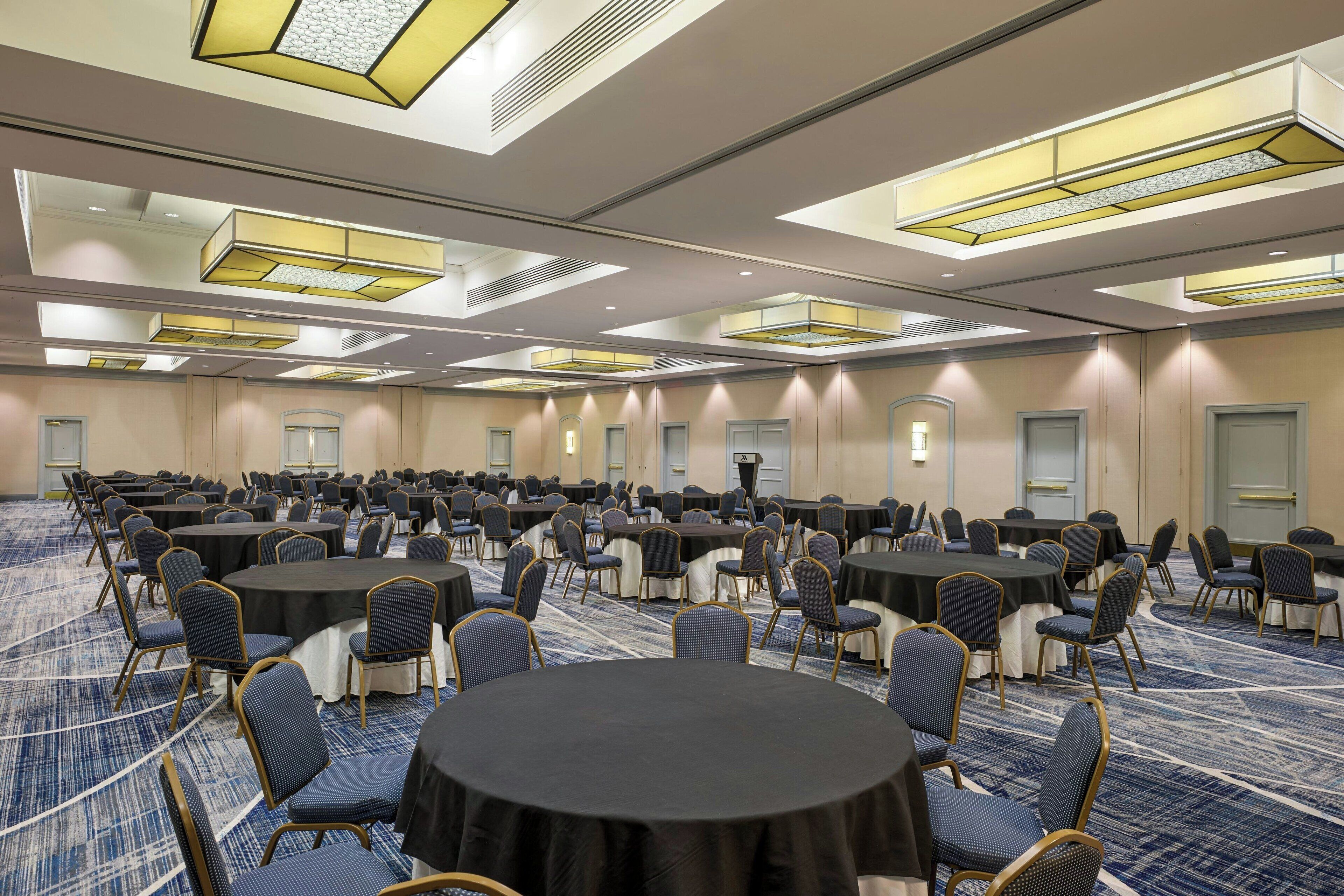 Een ruime congreszaal met meerdere ronde tafels en stoelen, uitgerust met plafondlampen en een vloerbedekking.