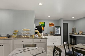 A modern kitchen with a marble countertop, a built-in oven, and a refrigerator. There are two dining tables with chairs, and a small island with a sink and stools.
