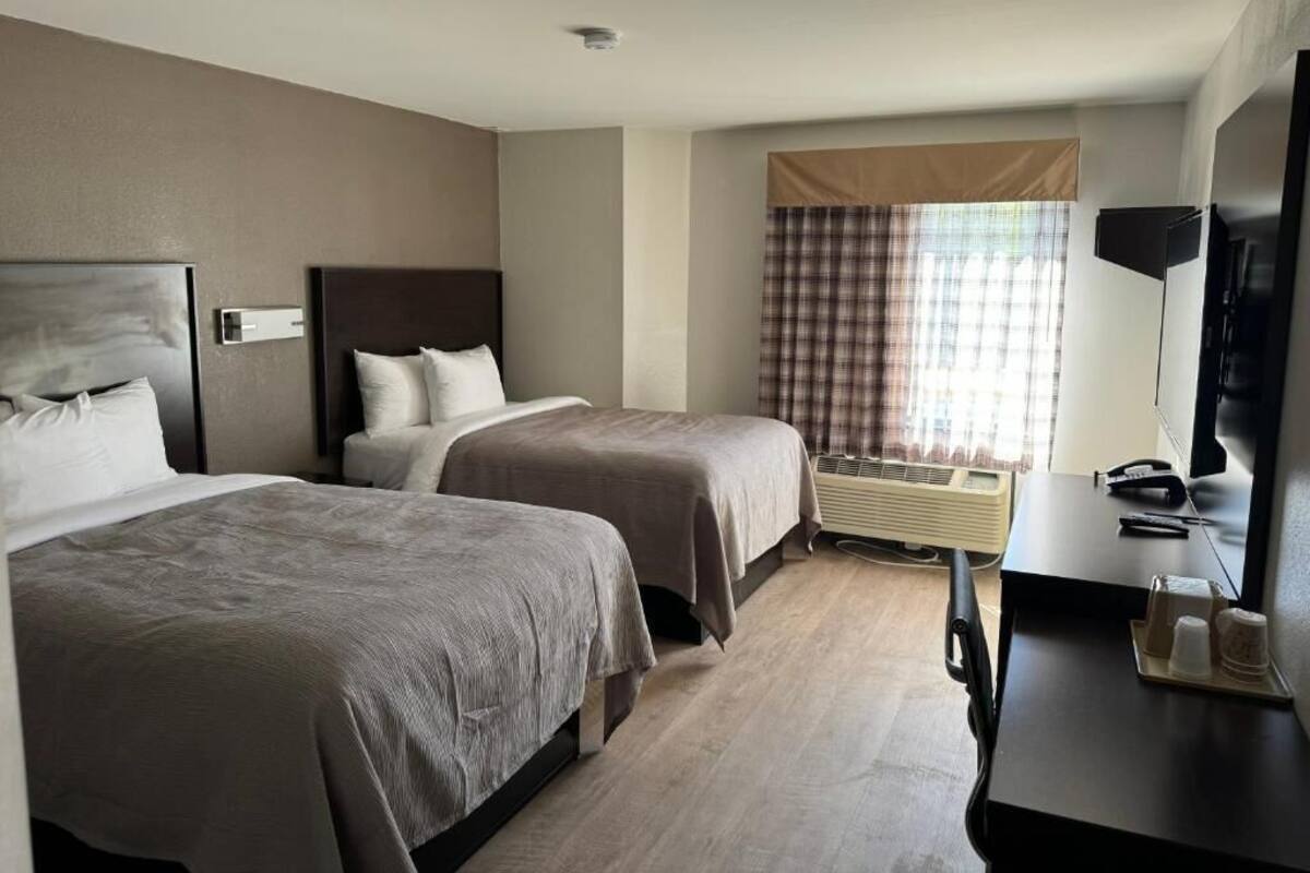 A hotel room with two beds, a desk, a flat-screen TV, and a window with curtains.