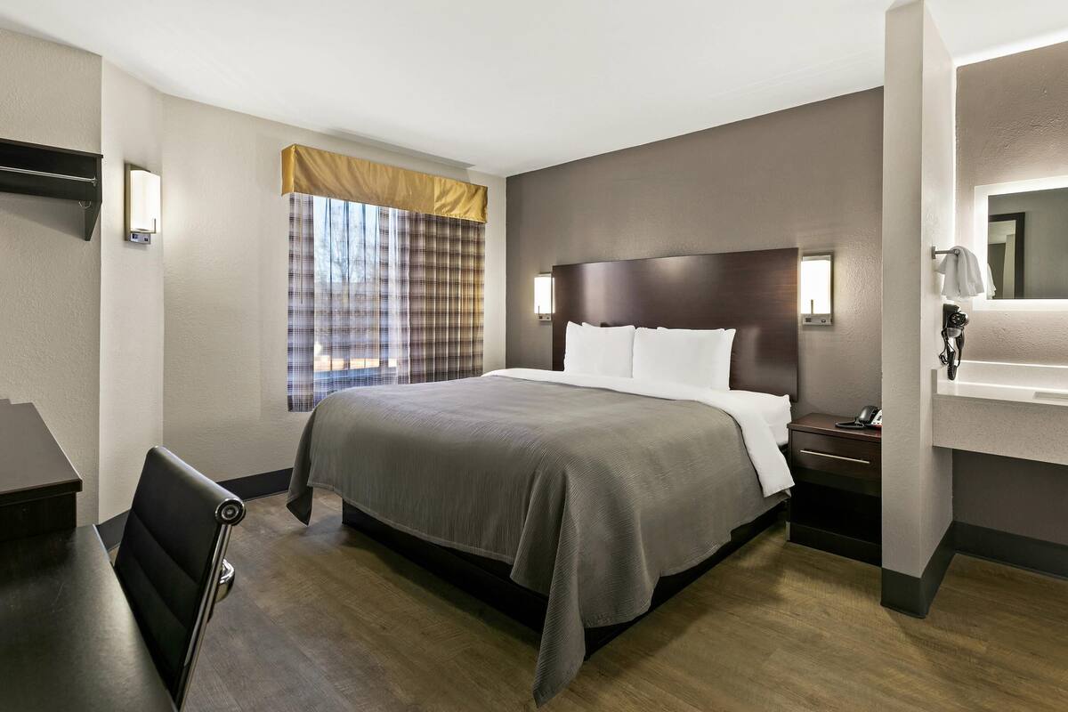 A hotel room with a large bed, a desk, a chair, a nightstand, a bedside table, a window with curtains, and a mirror.