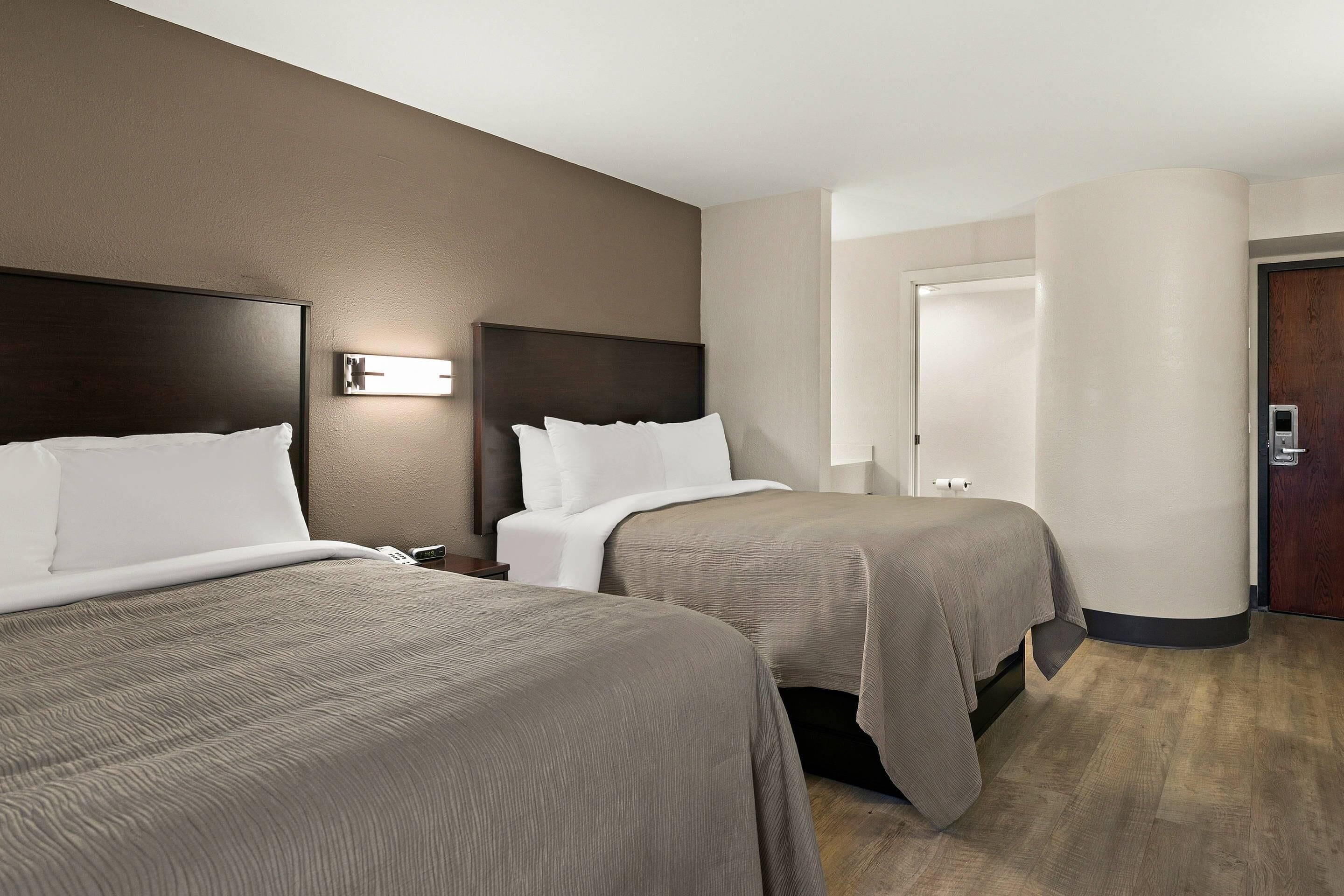 Habitación de hotel con dos camas, una mesita de noche, una lámpara de pared y una puerta.