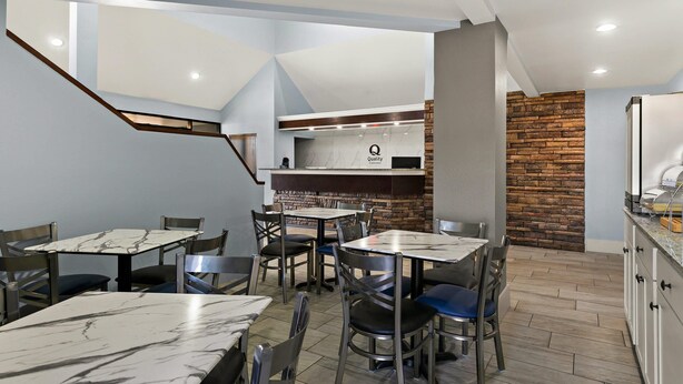 A modern dining area with marble tables and chairs, a kitchen area with a 'Quality' sign, and a brick accent wall. - Quality Inn Lavonia I-85 (Lavonia)