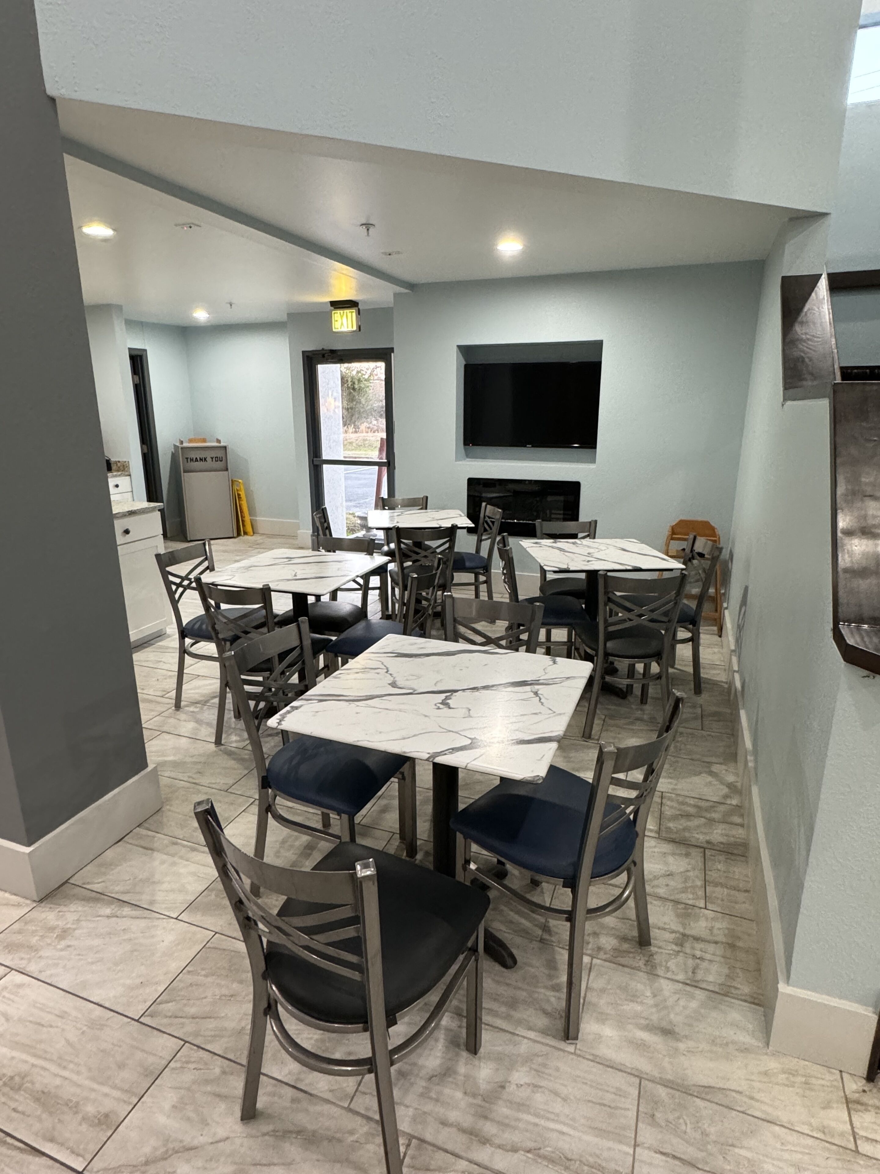 Un coin repas avec des tables et des chaises en marbre, un téléviseur à écran plat fixé au mur et une pancarte « Merci » sur un meuble.