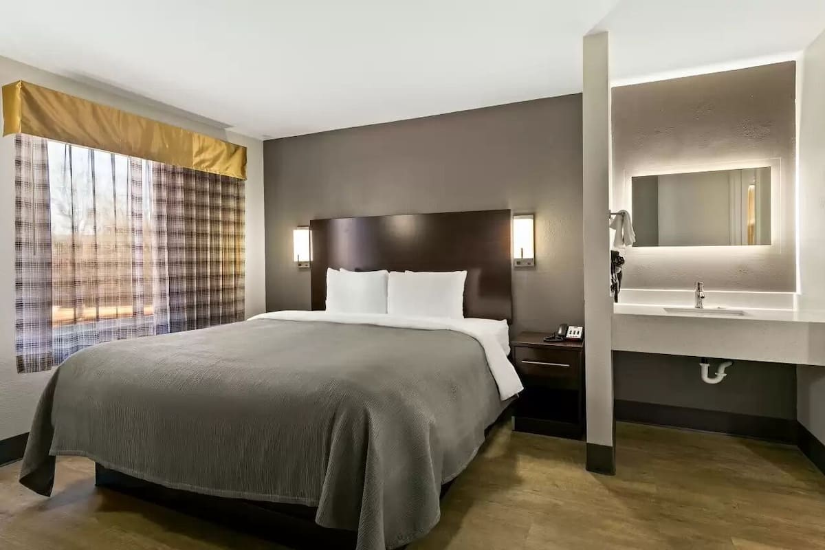 A hotel room with a bed, bedside table, nightstand, and a bathroom with a sink and mirror.