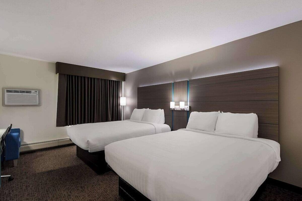 A hotel room with two beds, a desk, a chair, and a wall-mounted air conditioner.