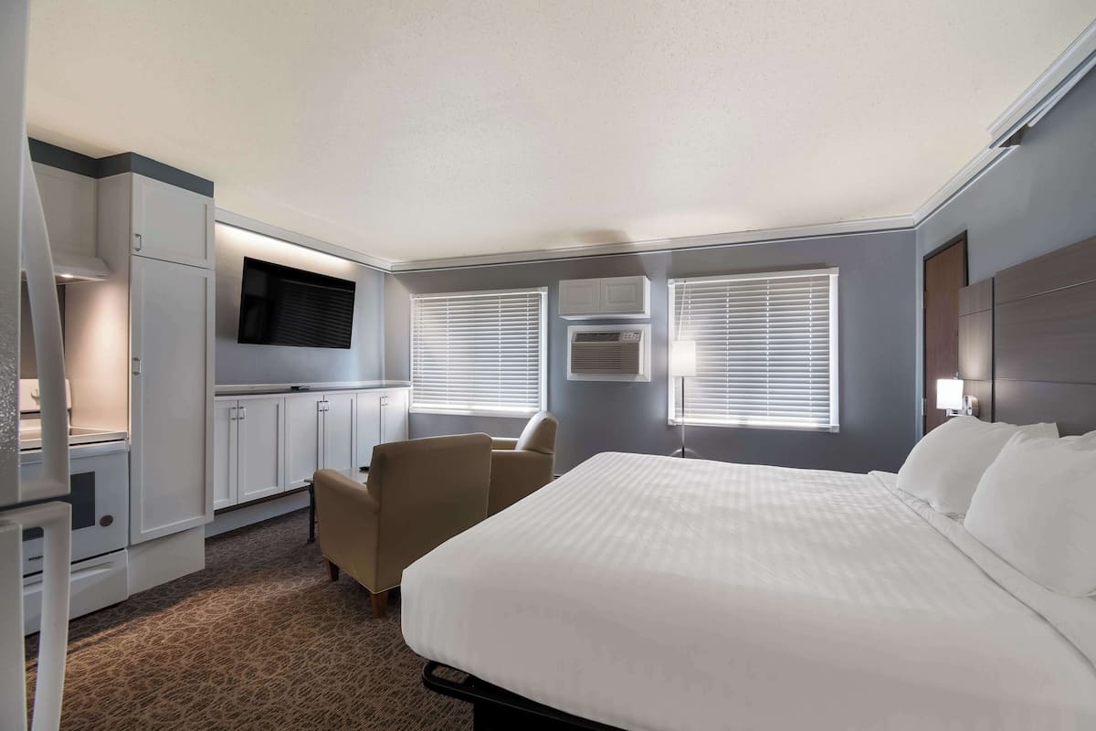 A hotel room with a bed, a desk, a chair, a TV, and a kitchenette.