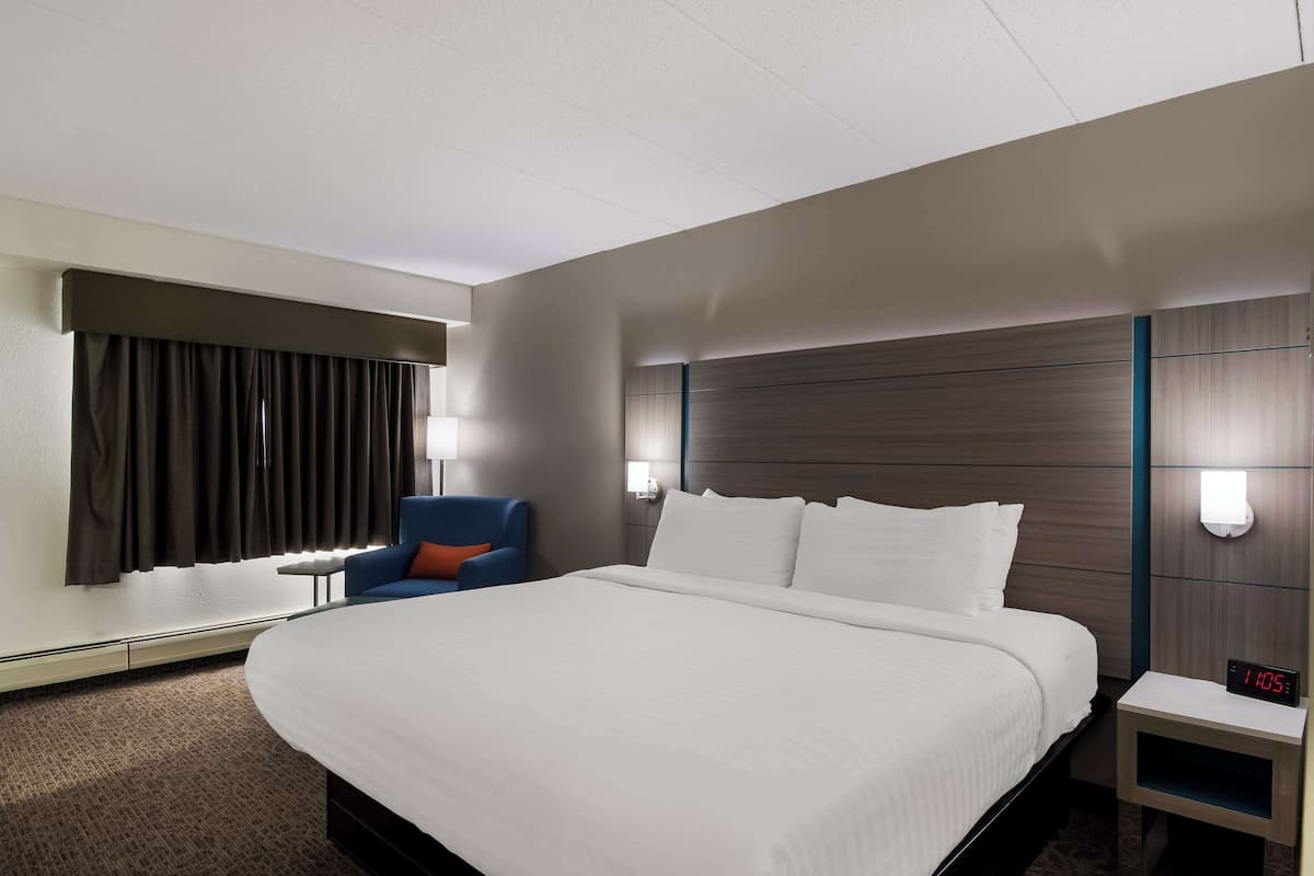 A modern hotel room with a large bed, a blue armchair, and a nightstand with a digital clock.