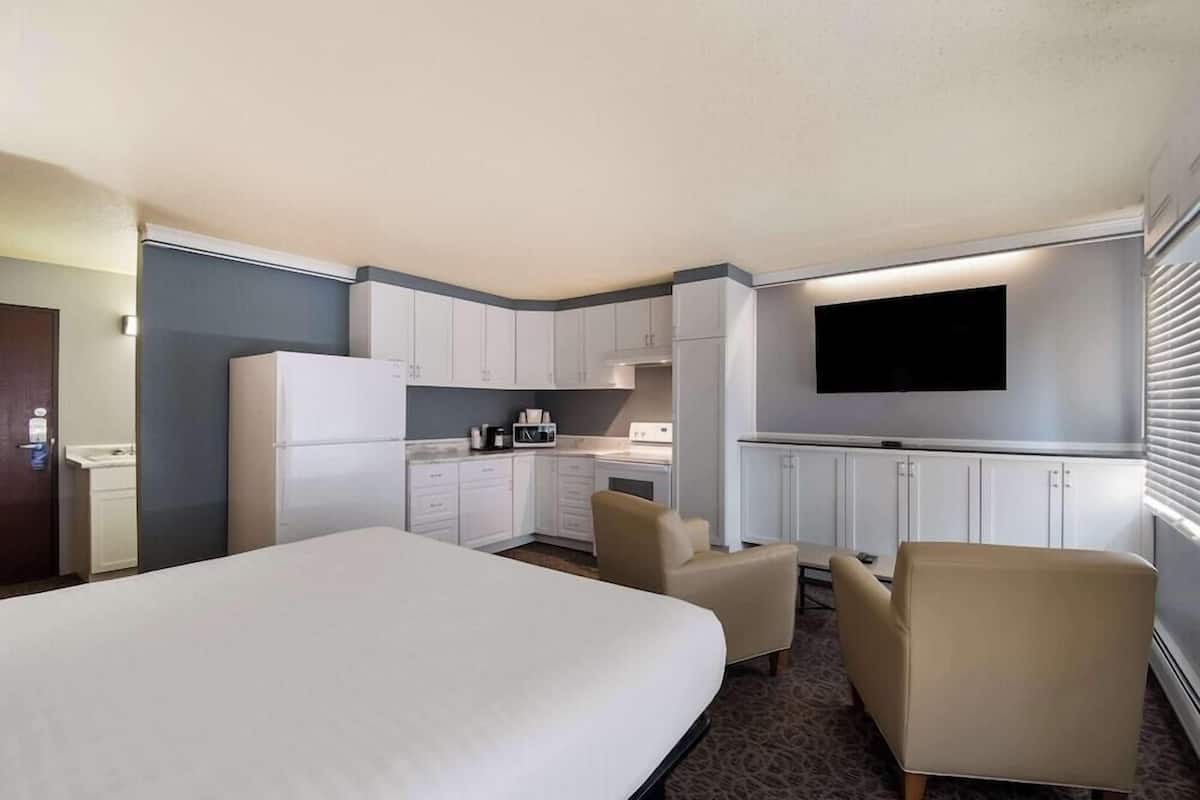 A hotel room with a bed, a kitchenette, a flat-screen TV, and two armchairs.