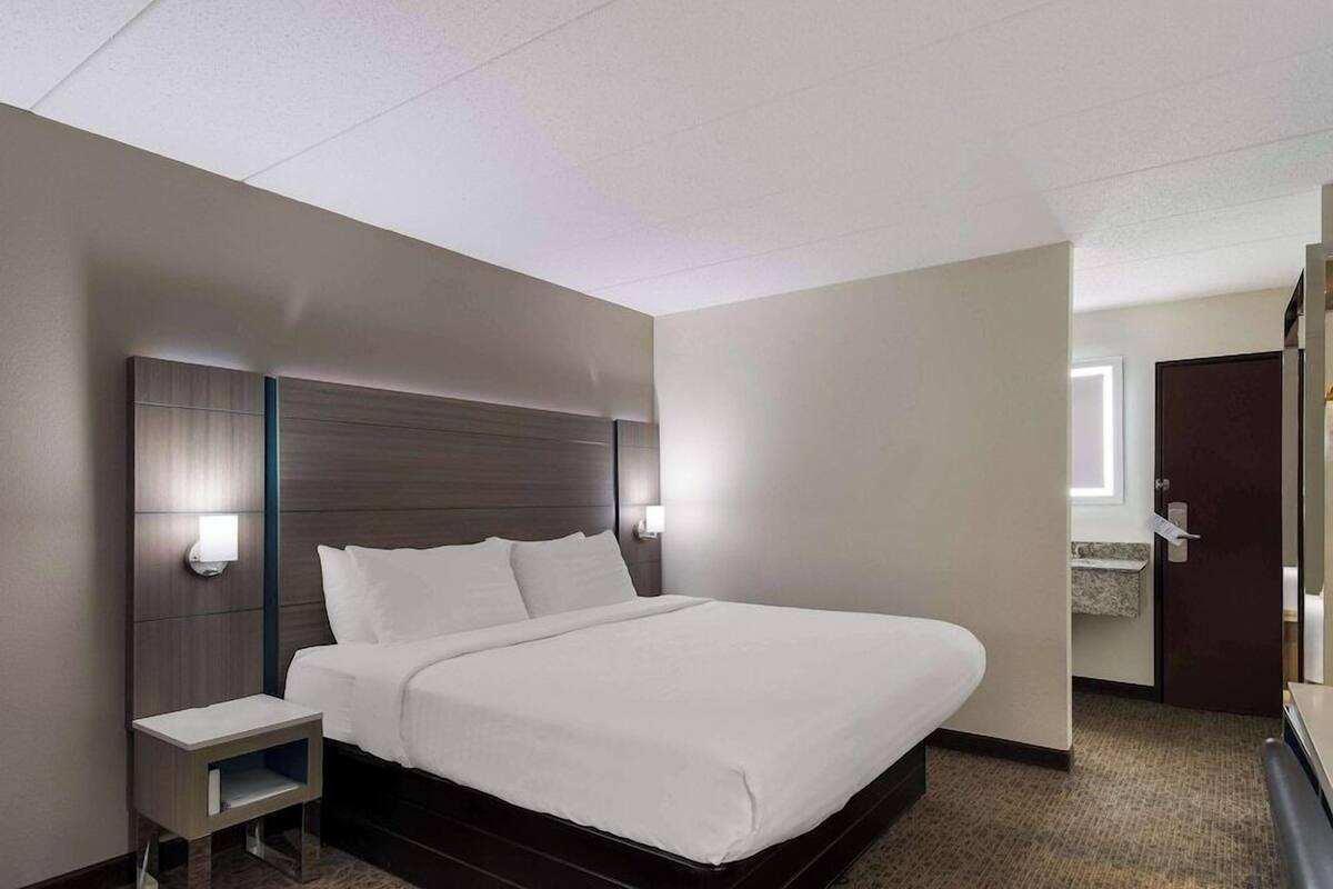 A hotel room with a large bed, bedside tables, a nightstand, a wall-mounted light, and a door leading to a bathroom.