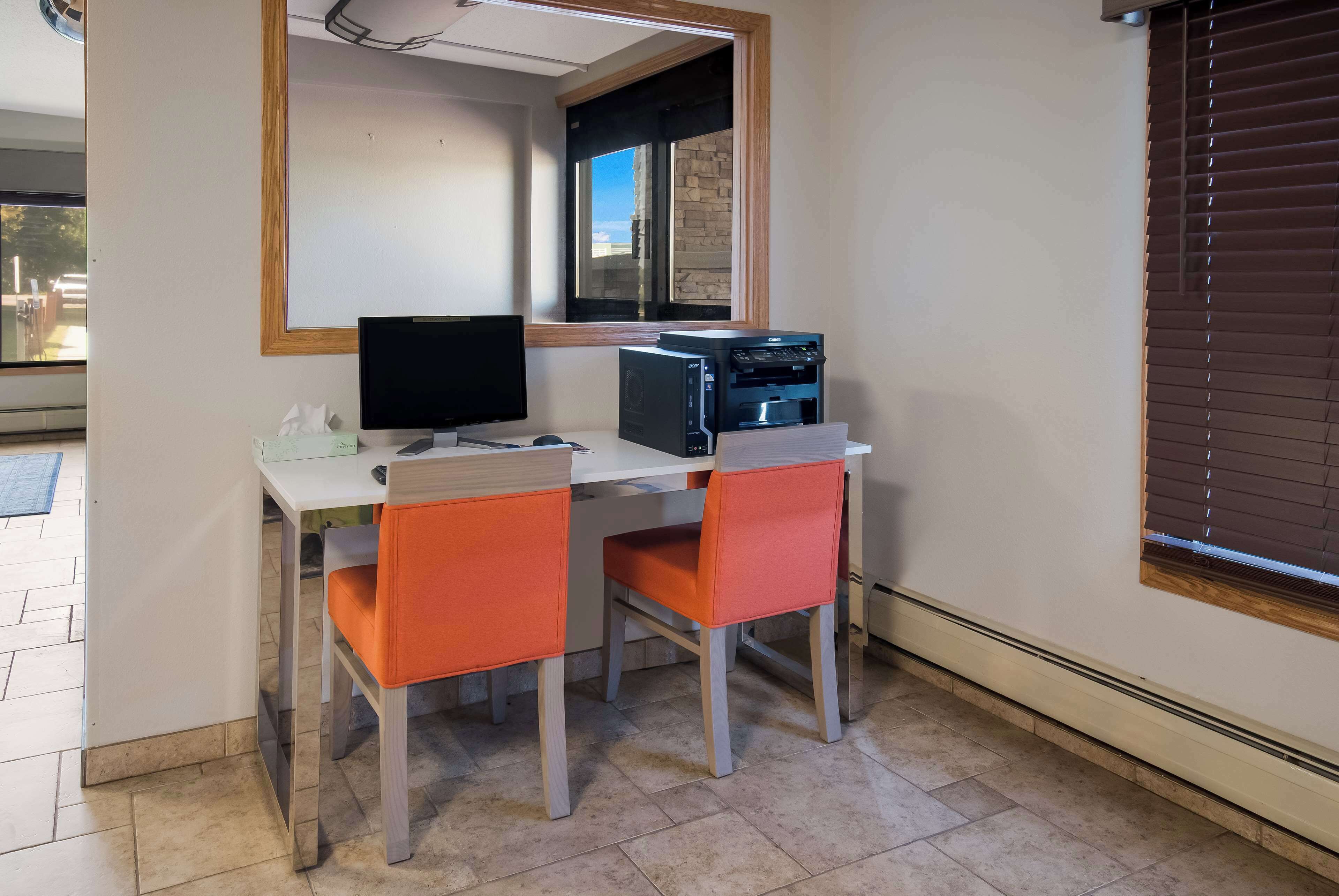 A small office with a desk, two chairs, a computer monitor, a printer, and a window with blinds.