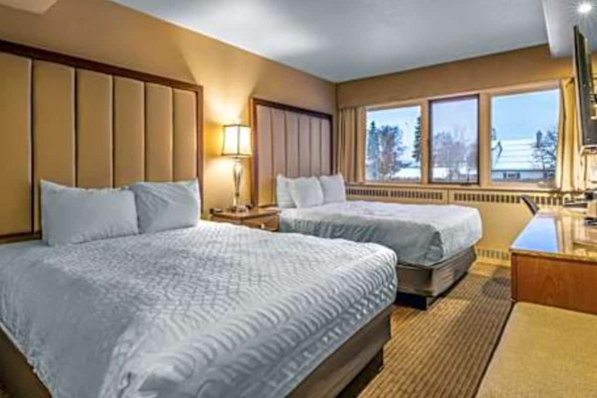 A hotel room with two beds, a window with a view, a desk, and a lamp.