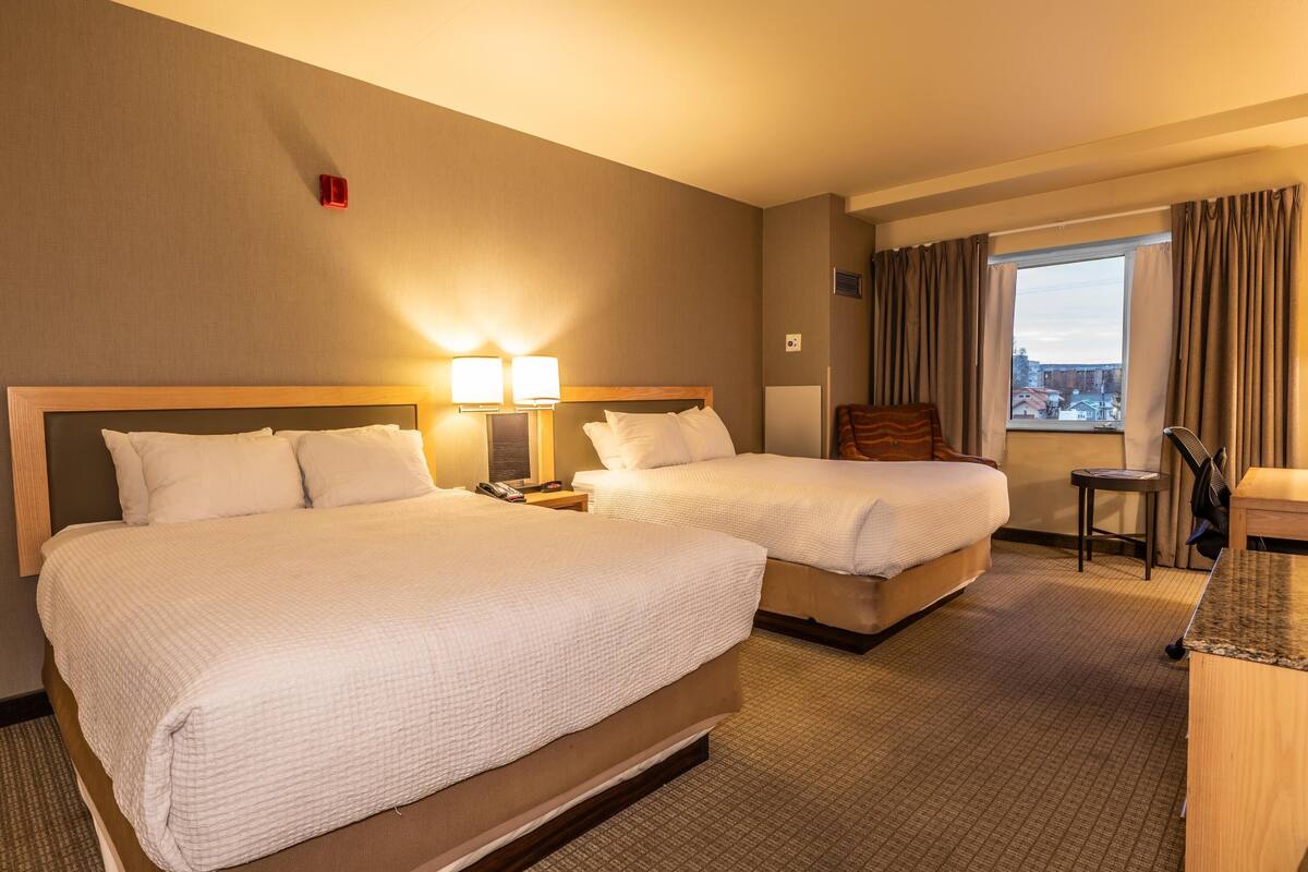 Une chambre d’hôtel avec deux lits, un bureau, une chaise, une fenêtre avec des rideaux et une vue sur les bâtiments environnants.