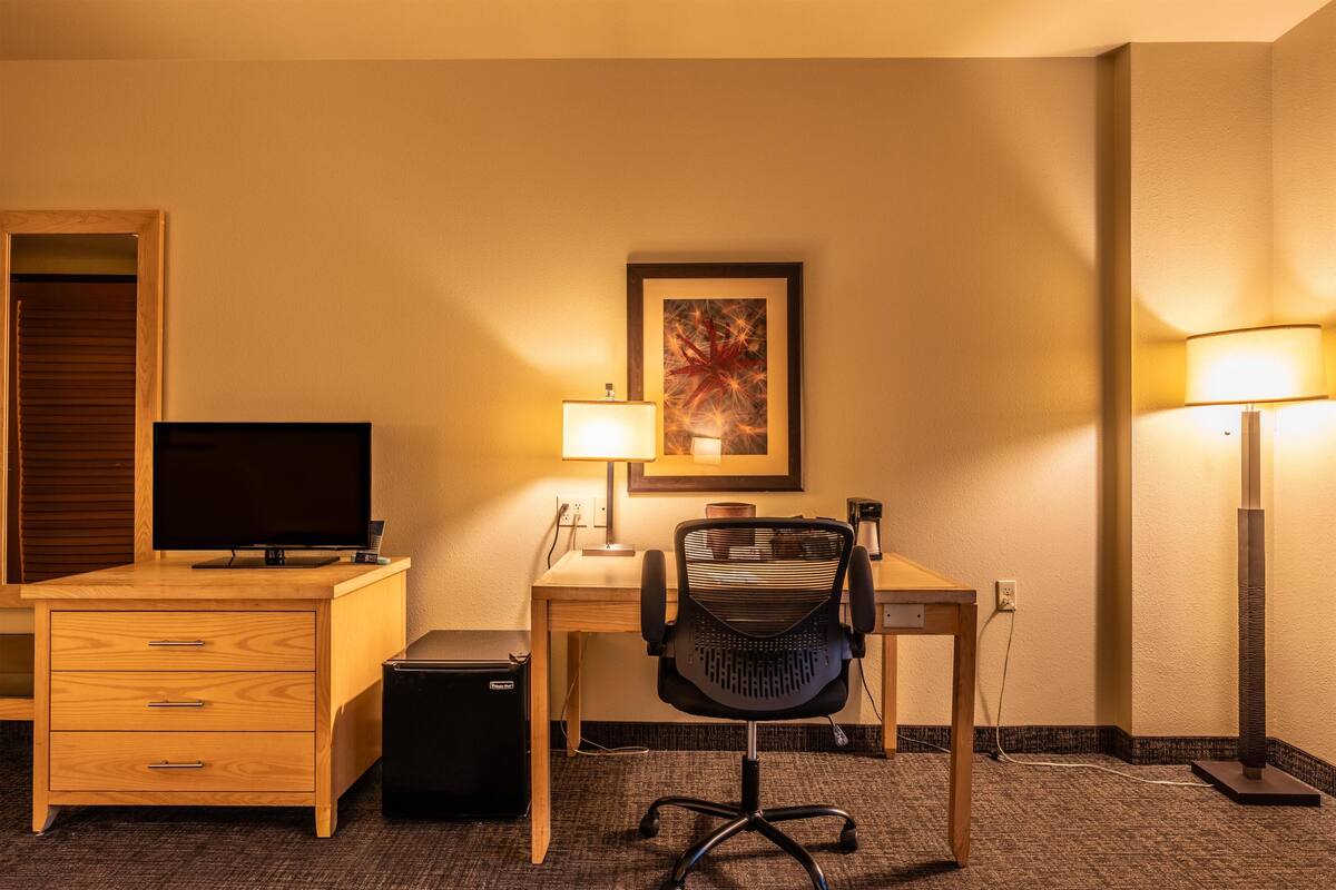 Une chambre d’hôtel avec un bureau en bois, un fauteuil de bureau noir, une télévision, une lampe et un tableau encadré au mur.