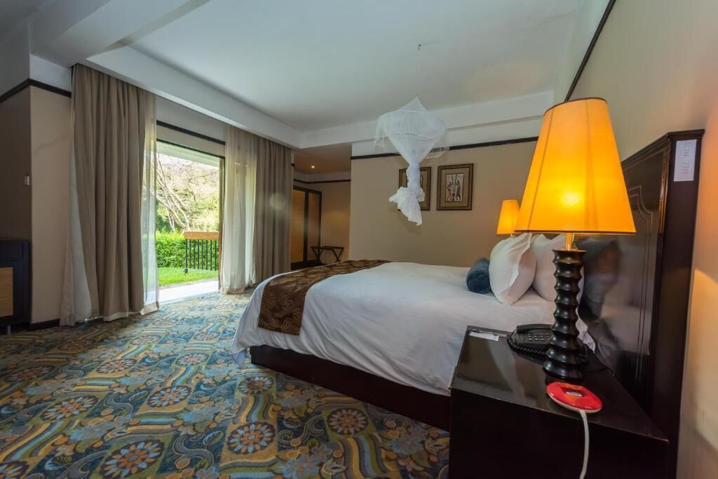 A hotel room with a bed, a bedside table with a lamp, a telephone, and a view of the outdoors through a large window.