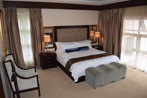 Una habitación de hotel con una cama grande, dos mesitas de noche con lámparas, una silla y un ventanal con cortinas.