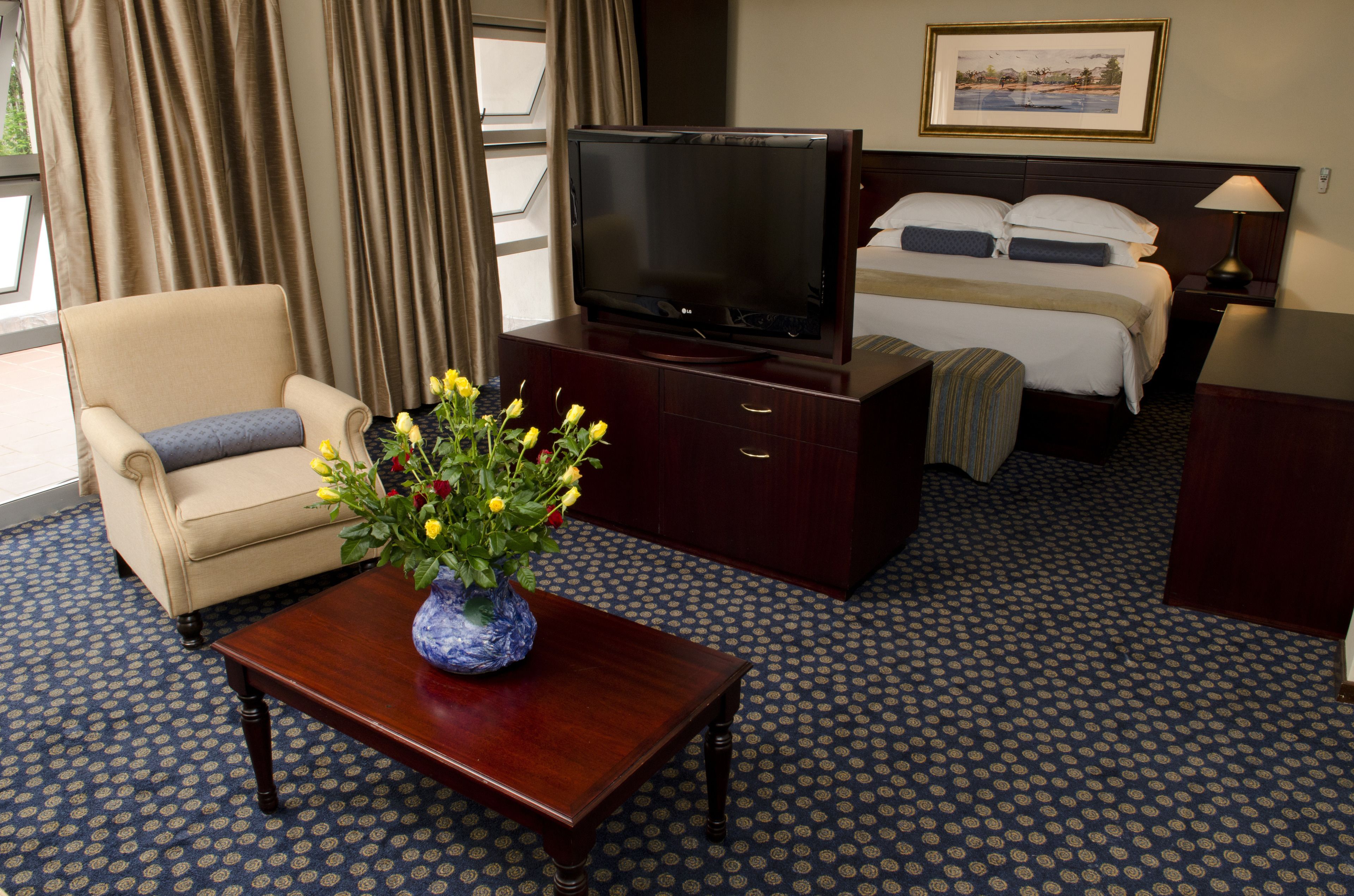 A hotel room with a bed, a TV, a desk with a vase of flowers, a chair, and a patterned carpet.